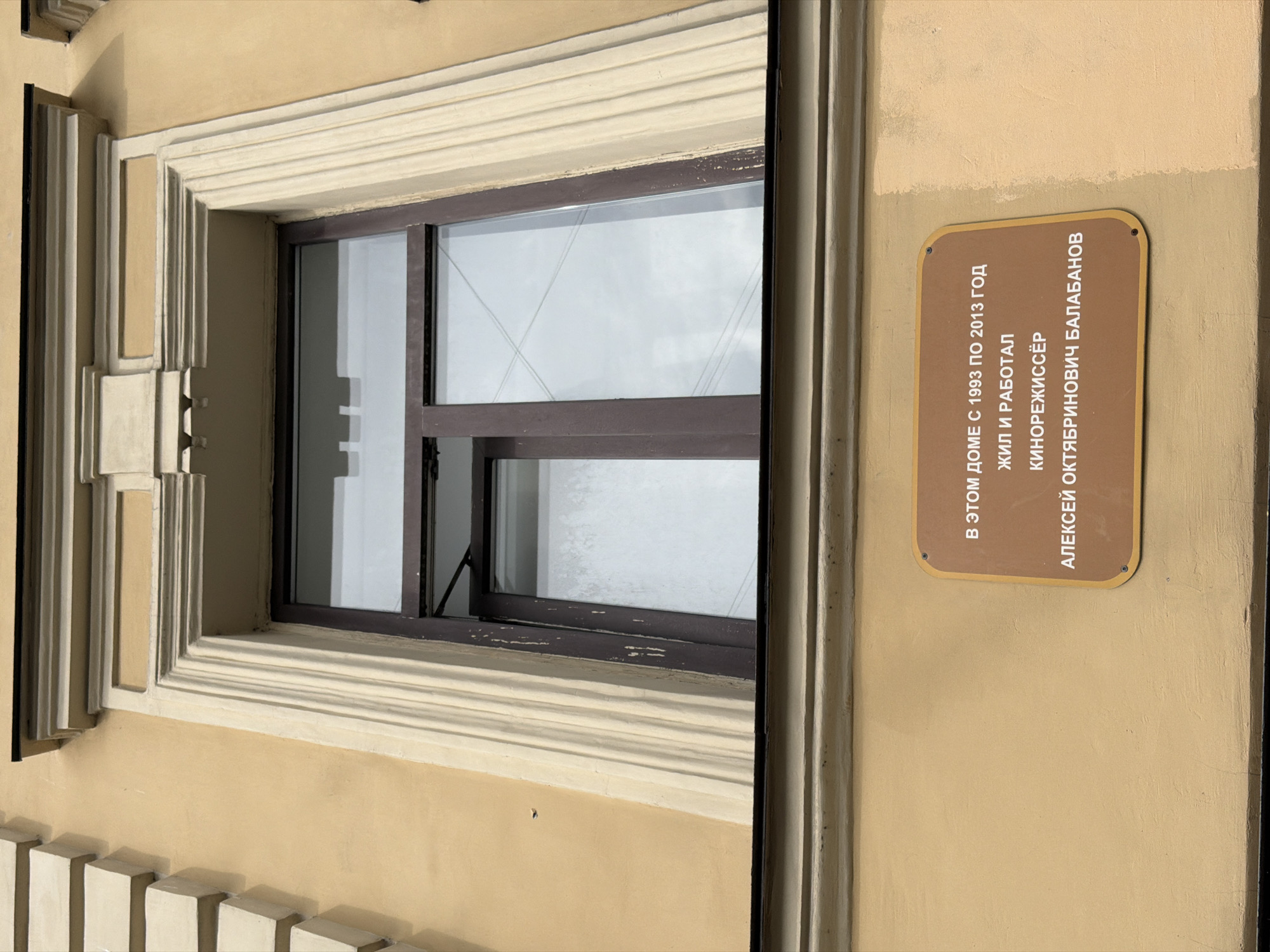 The image shows a memorial plaque on the exterior of a building in Saint Petersburg, Russia. The plaque is a light brown, rounded rectangle with dark brown text. It reads: "В ЭТОМ ДОМЕ С 1993 ПО 2013 ГОД ЖИЛ И РАБОТАЛ КИНОРЕЖИССЁР АЛЕКСЕЙ ОКТЯБРИНОВИЧ БАЛАБАНОВ". This translates to "In this house from 1993 to 2013 lived and worked film director Aleksei Oktyabrinovich Balabanov". The building appears to be yellow with ornate architectural details around a window. The window itself is a dark brown frame with multiple panes, reflecting an overcast sky. The plaque is mounted on the lower portion of the building's facade.