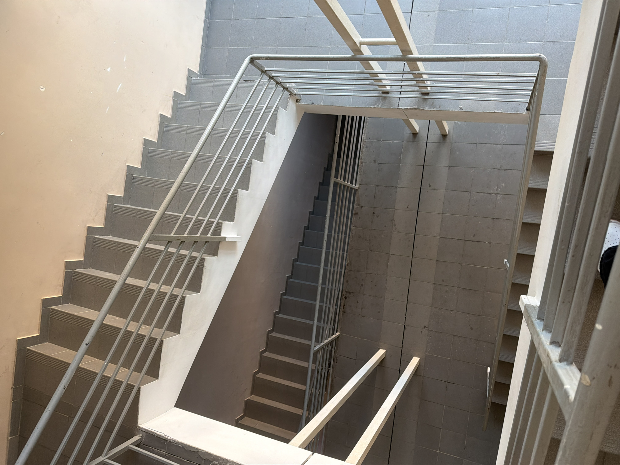 This media file depicts an interior view of a concrete staircase with metal railings. The scene is well-lit, suggesting it could be daytime. The staircase is constructed with grey tiled steps and a light-colored wall on one side, contrasted with dark grey tiled walls on the other. Metal railings with vertical bars line the staircases, providing safety and a sense of depth to the composition.
The perspective is from an elevated position, looking down the stairwell. There are two distinct staircases visible, one on the left and another on the right, with a landing area between them. The staircase on the left ascends, while the one on the right descends. White beams are visible spanning across the top of the stairwell, possibly part of the building's structure. The overall impression is one of utilitarian architecture, common in institutional or residential buildings. The location context of Kyiv, Ukraine, implies a potentially urban or suburban setting. No individuals or text are visible in the image.