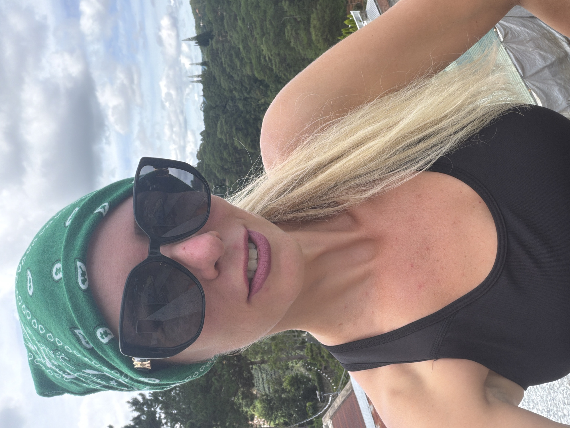 A blonde woman, wearing a green bandana and oversized black sunglasses, is captured in a close-up selfie. She has on a black sports bra. Her mouth is slightly open, revealing pink lipstick. Her blonde hair is pulled back and falls over her shoulder. The background is a lush, green hillside with scattered trees and some buildings, suggesting an outdoor location in a hilly or mountainous area. In the distance, several tall, conical trees are visible against a cloudy sky. The lighting suggests it is daytime. The context of Da Lat, Vietnam, is provided, implying this scene is set within the city's known scenic and verdant landscape.