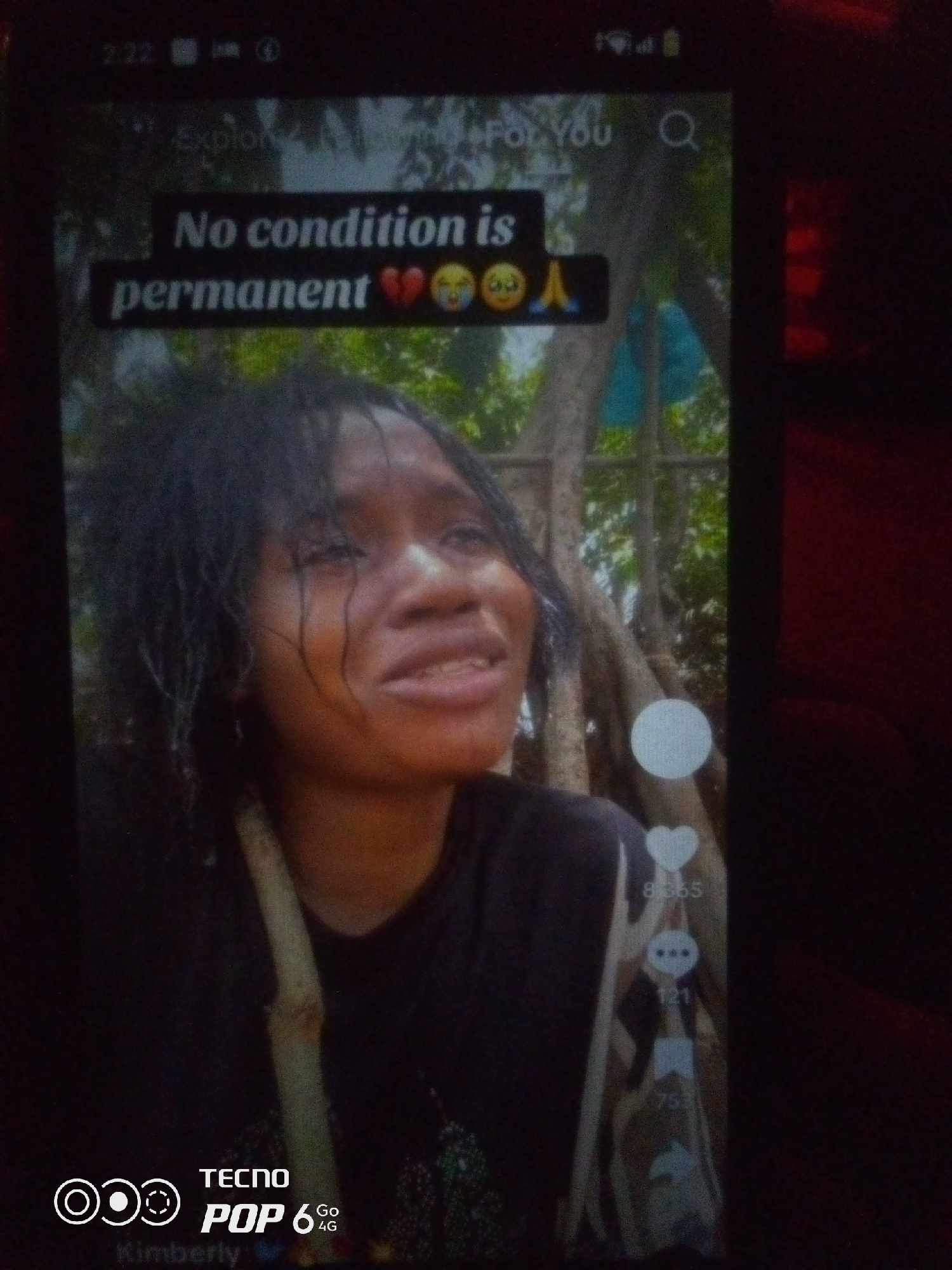 The image displays a screenshot of a social media video on a phone screen. A young Black woman with wet, dark hair is the central subject. She appears to be crying, with tears streaming down her face and a distressed expression. Her mouth is slightly open, and she is wearing a dark t-shirt. Overlayed on the video is text that reads "No condition is permanent" along with emojis of a broken heart, a crying face, and praying hands. The background of the video appears to be an outdoor setting with trees and some foliage. On the right side of the phone screen, social media interaction icons are visible, including a heart with the number 8,865, a speech bubble with the number 121, and a share icon with the number 753. In the bottom left corner of the phone screen, the TECNO POP 6 4G logo and the name "Kimberly" are visible. The surrounding environment of the phone is dark and appears to be indoors. There is no discernible information within the image that places it in the city of Howayi, Nigeria.