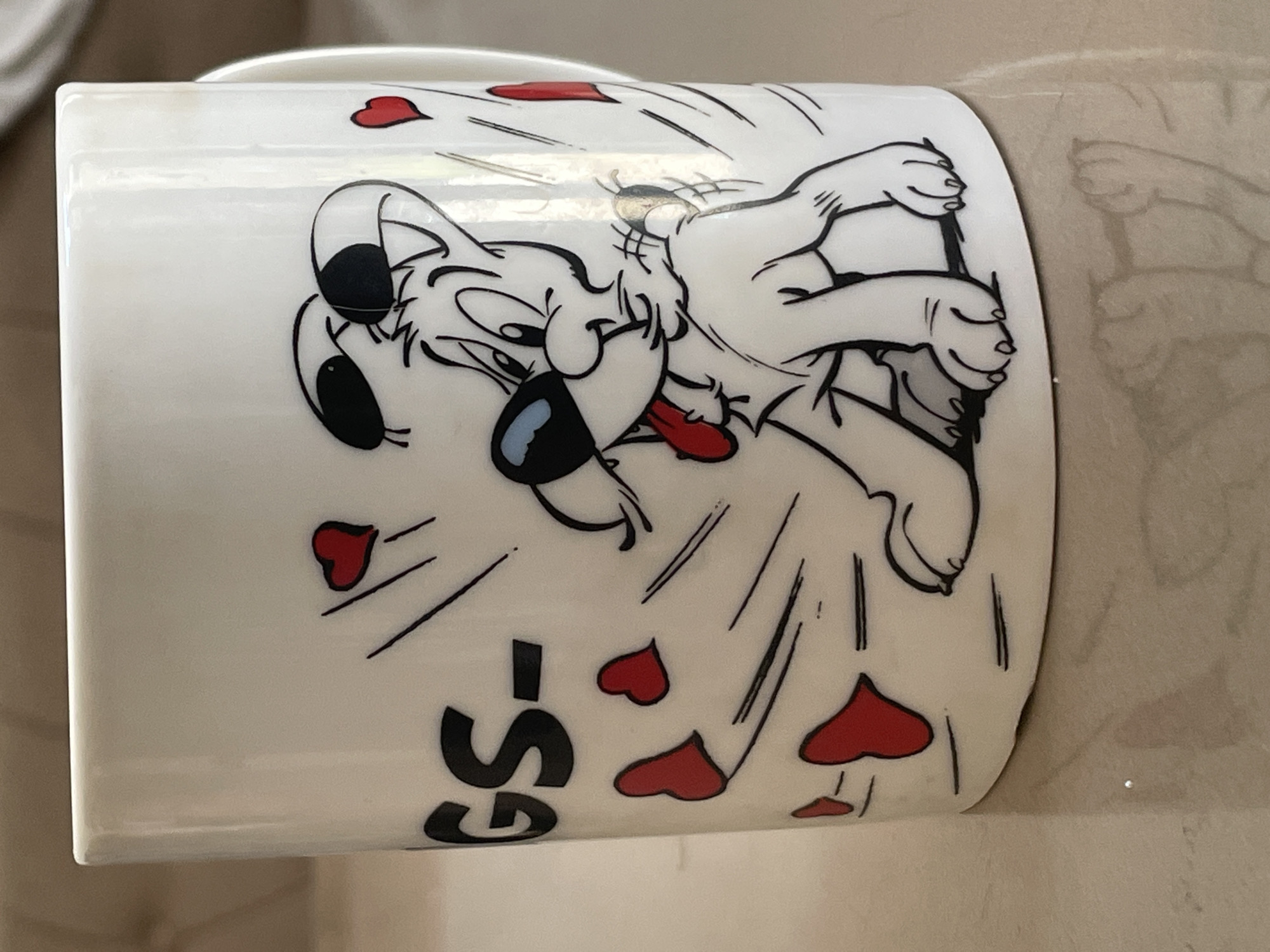 A white ceramic mug with a cartoon dog and red hearts is displayed on a reflective, light-colored surface, possibly a table. The mug features a black outline drawing of a white dog with black accents on its ears and eyes, and a red tongue sticking out. Several red hearts are scattered around the dog, with some appearing to be in motion. The word "GS-" is visible in large black lettering on the left side of the mug. The mug is partially obscured by a handle on the right side. The background is out of focus, showing a textured surface. There is no indication of the city of Tashkent, Uzbekistan, or any urban environment.