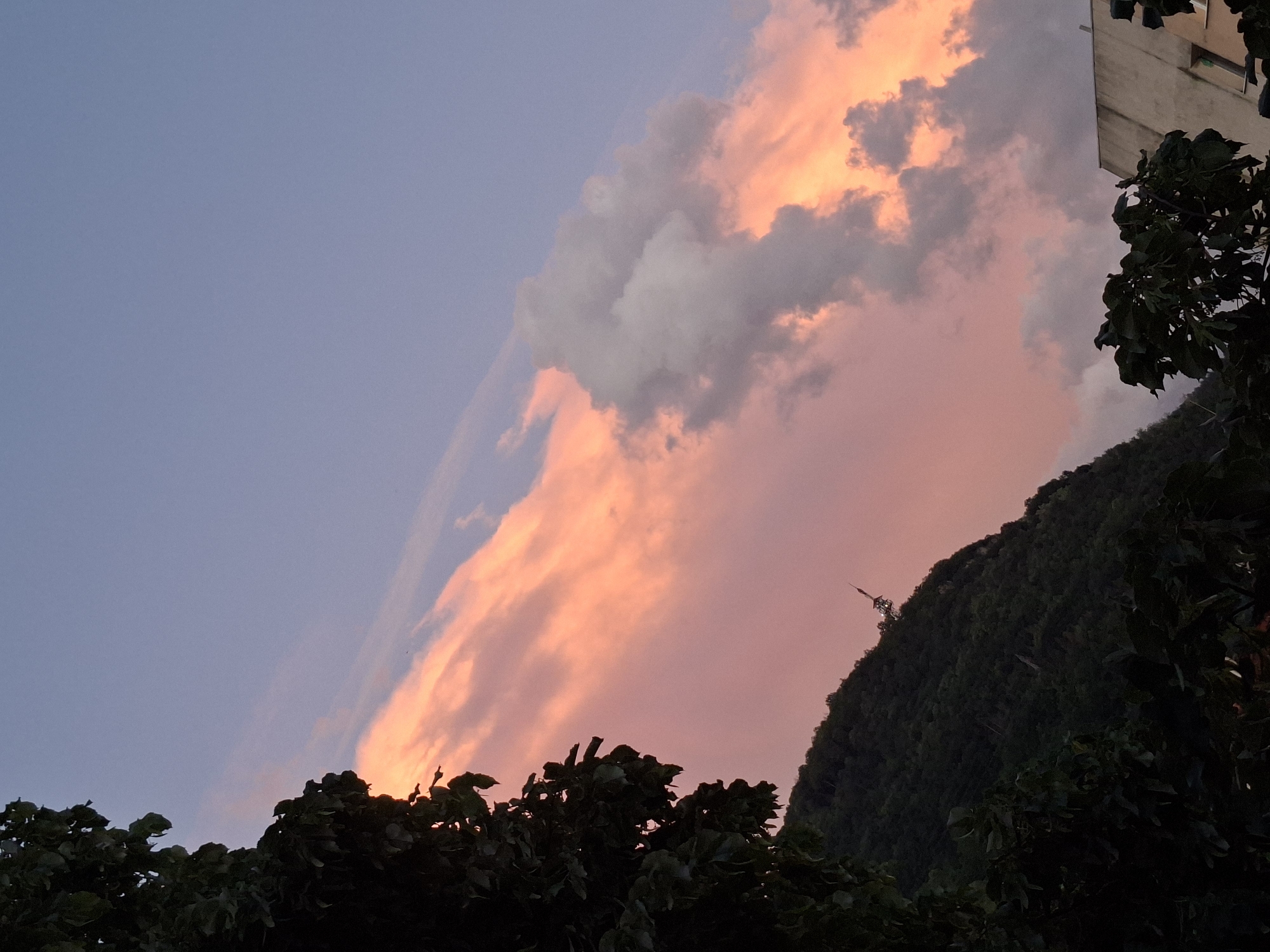 Captured from Gabrovo, Bulgaria, the image presents a dusk scene dominated by colorful clouds. The sky transitions from a pale blue in the upper-left to a vibrant blend of pinks and oranges in the center and right, where the clouds are concentrated. These clouds vary from fluffy white to darker gray hues. A mountain densely covered with green foliage is visible on the right side of the frame, with a communications tower perched atop its peak. In the foreground, the silhouettes of treetops add a dark, textural border to the bottom edge of the image. A building can be partially seen in the upper-right corner.