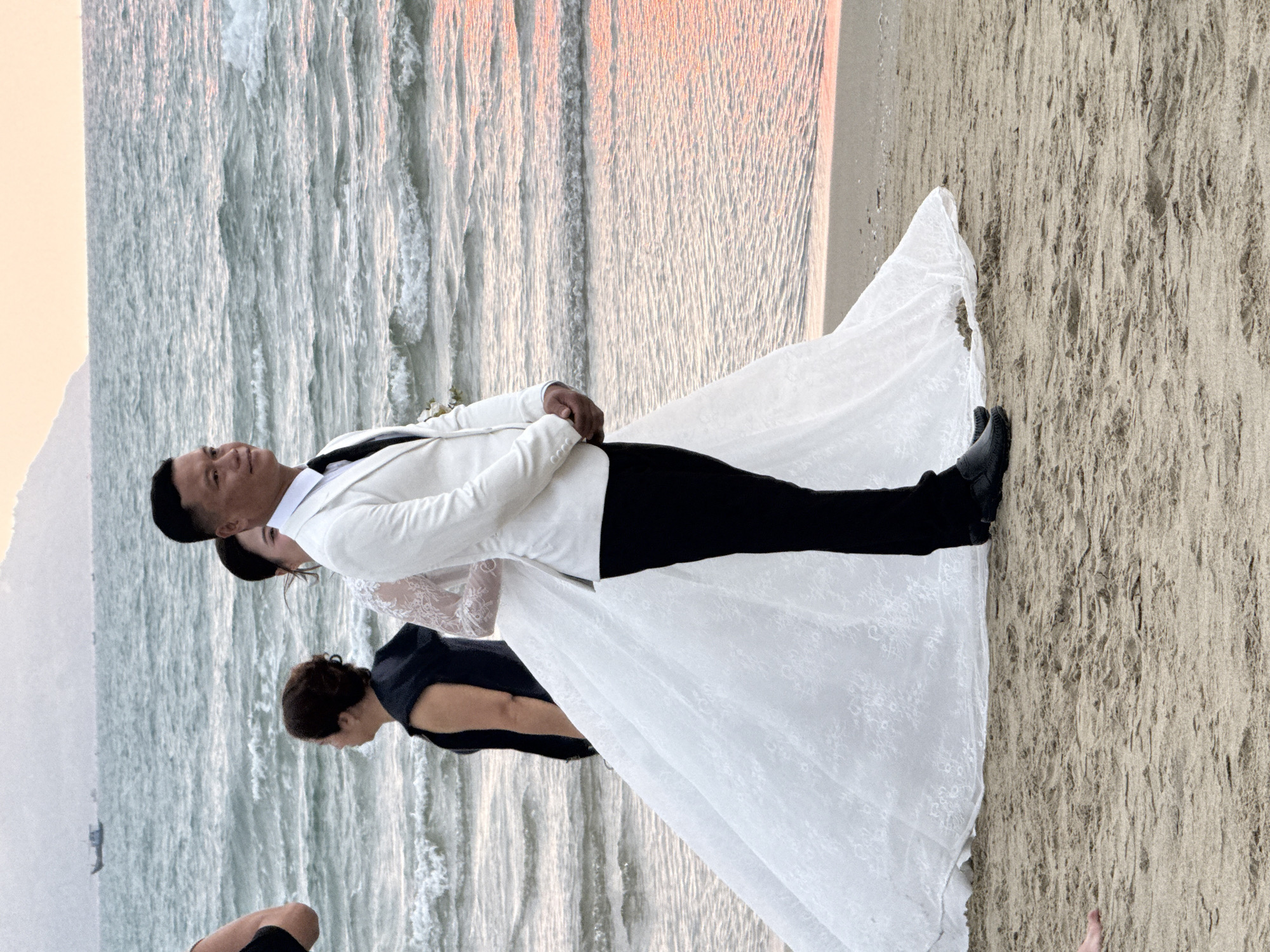 A wedding photo is taken on a beach in Đà Nẵng, Vietnam, at sunset. The bride and groom are embracing, standing on the sand. The groom is wearing a white suit jacket with black pants and shoes. The bride is wearing a white lace wedding dress. The ocean and beach stretch out behind them. A woman is nearby, looking towards the bride and groom. The sky in the background is colored with a mixture of orange and pink hues.