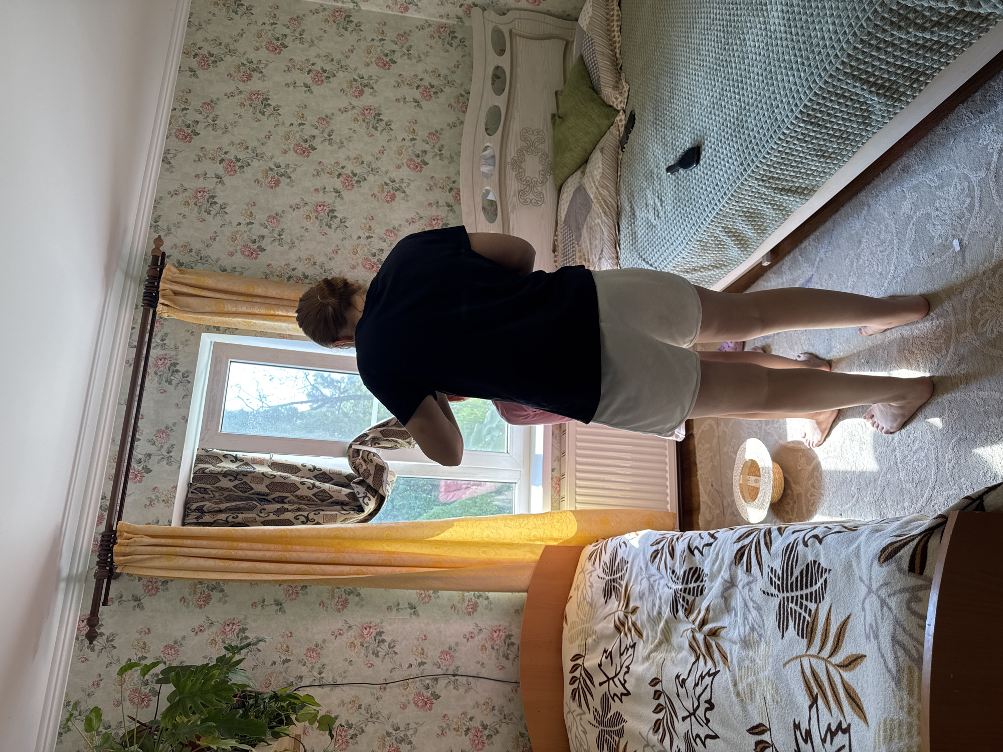 The media file depicts an indoor scene within a bedroom in Zassovskaya, Russia. The room is decorated with floral wallpaper, featuring a pattern of pink and green flowers. Two beds are visible; one is partially covered with a light green textured blanket, and the other is mostly covered by a cream-colored blanket with a brown leaf pattern. A white headboard with decorative cutouts is positioned between the two beds.
A person, seen from the back and side, is standing and appears to be leaning over. They are wearing a dark blue t-shirt and white shorts. Their hair is tied back. Another person, a child, is partially visible beneath the standing person, wearing pink clothing and lying down.
To the left of the standing person, a window is open, allowing sunlight to stream into the room, indicating it is daytime. Yellow curtains are drawn on either side of the window. Outside the window, trees and foliage are visible, suggesting a residential or natural setting. A straw hat is placed on the light-colored carpet near the standing person's feet.
The overall impression is a candid, everyday moment captured in a domestic environment. The bright sunlight and the casual attire suggest a warm day, possibly during the summer.