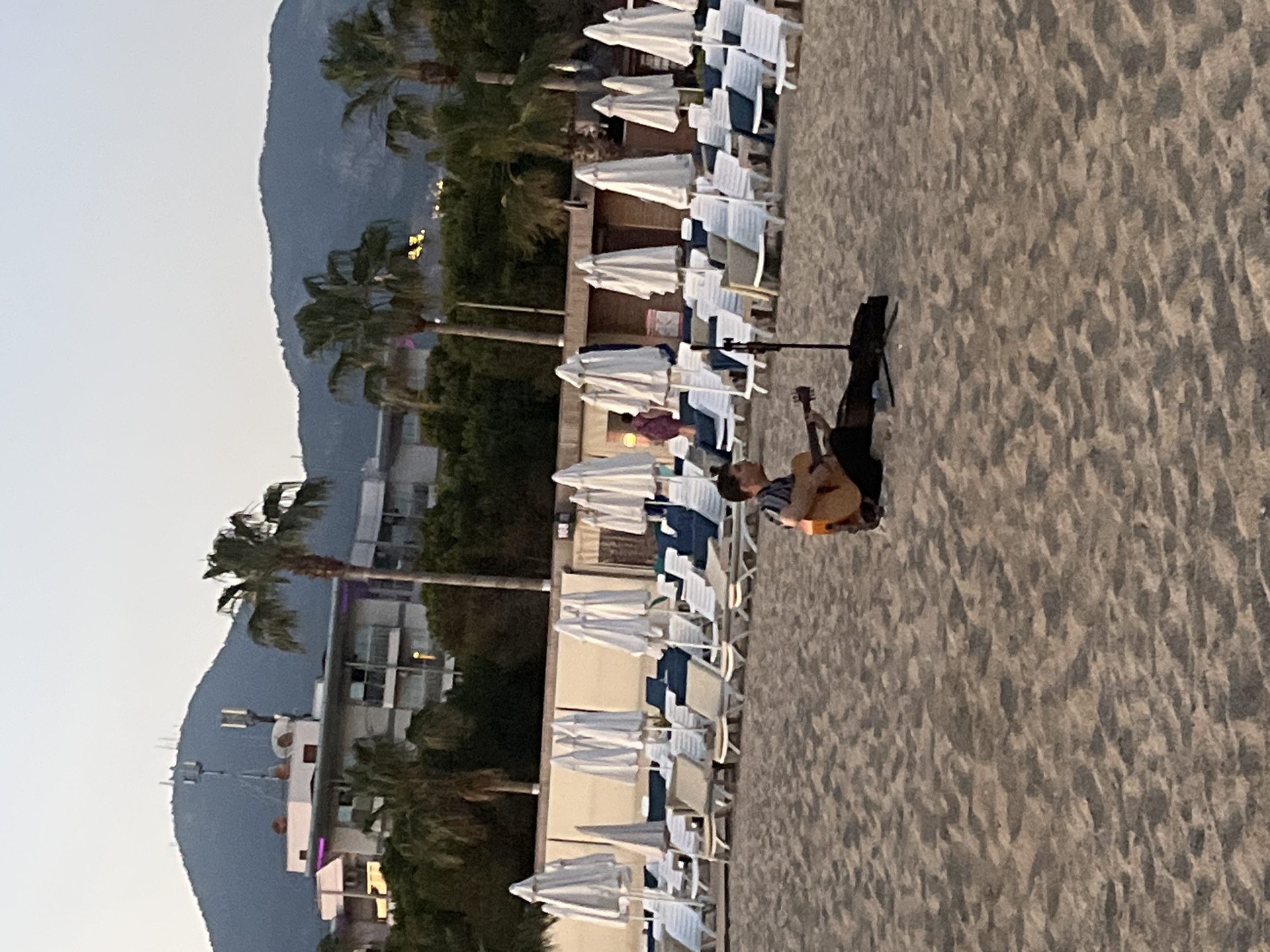 The image depicts a beach scene in Alanya, Turkey. A man is seen playing a guitar on the sandy beach. Rows of white beach chairs with blue accents are lined up neatly behind him, with white umbrellas placed next to each chair. Palm trees are interspersed between the chairs, and in the background, there is a building. Mountains are visible in the distance, and the sky is a pale blue.