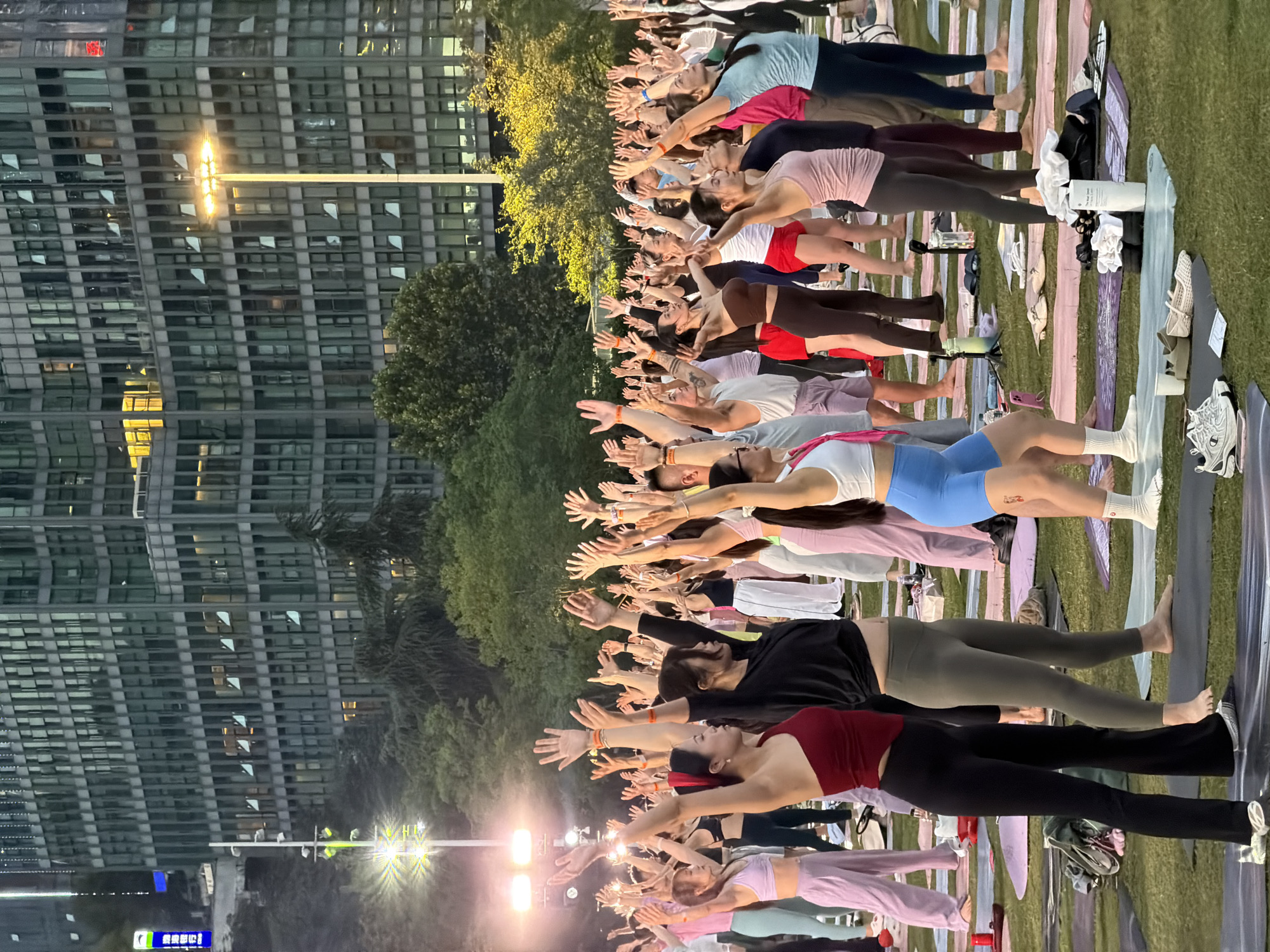 An outdoor yoga class is taking place on a grassy area in front of a modern, glass-fronted building in the city of Jiangbeicheng Subdistrict, China. A large group of people, primarily women, are standing on yoga mats with their arms raised in the air, seemingly in unison. They are dressed in athletic wear, with various colors and styles. The building behind them has many windows, some illuminated with warm light, suggesting it is a commercial or office space. Trees are visible between the building and the yoga participants. Streetlights are also present, casting a bright glow on the scene. The overall atmosphere appears to be one of communal wellness activity in an urban setting.