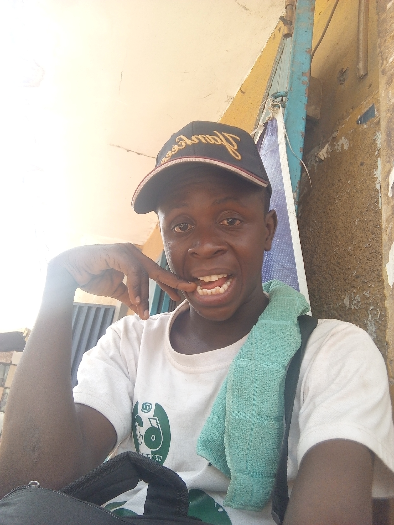 A young person, appearing to be male with dark skin, is captured in a bright, outdoor selfie. He wears a black baseball cap with gold cursive text that reads "Pancakes" (reversed due to the selfie perspective). His expression is energetic, with wide eyes and an open mouth revealing his teeth, suggesting a smile or playful shout. His right hand is raised, with the index finger near his mouth.
He is dressed in a white t-shirt featuring a circular green logo on the front, which appears to depict a stylized animal head, possibly a panda. A light blue, textured towel is draped over his left shoulder, and a black bag strap is visible across his chest and shoulder.
The background shows an outdoor setting, likely in Maiburiji, Nigeria, with bright, harsh sunlight illuminating the scene from the upper left, causing some overexposure. Behind him is a worn wall, painted in shades of yellow and brown, with peeling plaster. A prominent bright blue vertical structure, possibly a metal door frame or beam, runs vertically behind his right side. To the far left, a darker, possibly corrugated metal or shuttered structure is visible. The overall impression is casual and vibrant, taken in a sunlit, possibly urban or semi-urban environment.
