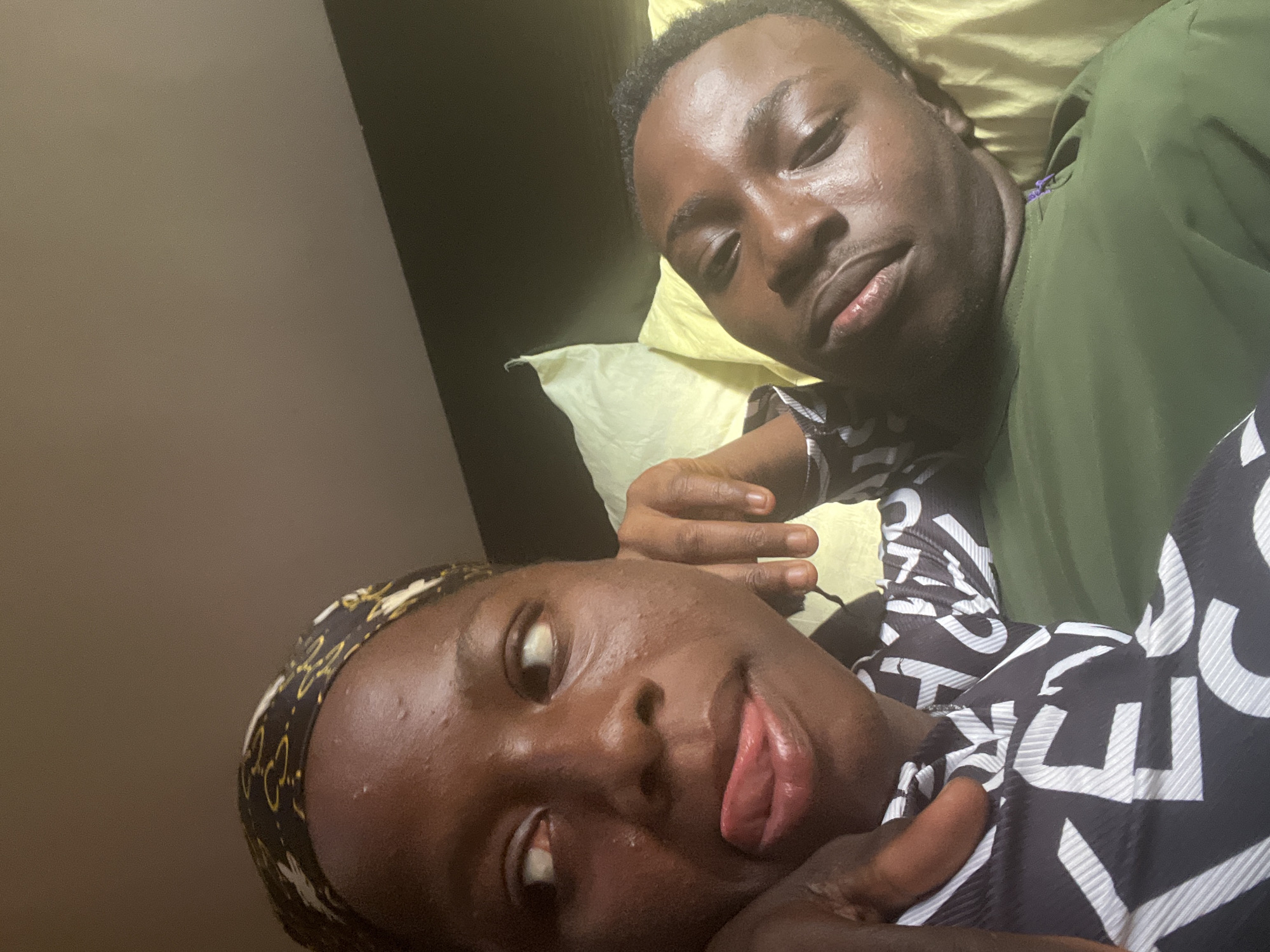 This image is a close-up selfie of a young African couple lying down in what appears to be a bedroom. The female subject on the left is wearing a black and gold patterned headscarf and a black and white patterned shirt. She is looking to her right with her tongue sticking out. The male subject on the right is wearing a green t-shirt and is lying on his side, looking towards the camera. They are both resting on a yellow pillow. The background is dimly lit and suggests an indoor environment, possibly a bedroom. Based on the context provided, the location is Nkpor, Nigeria.