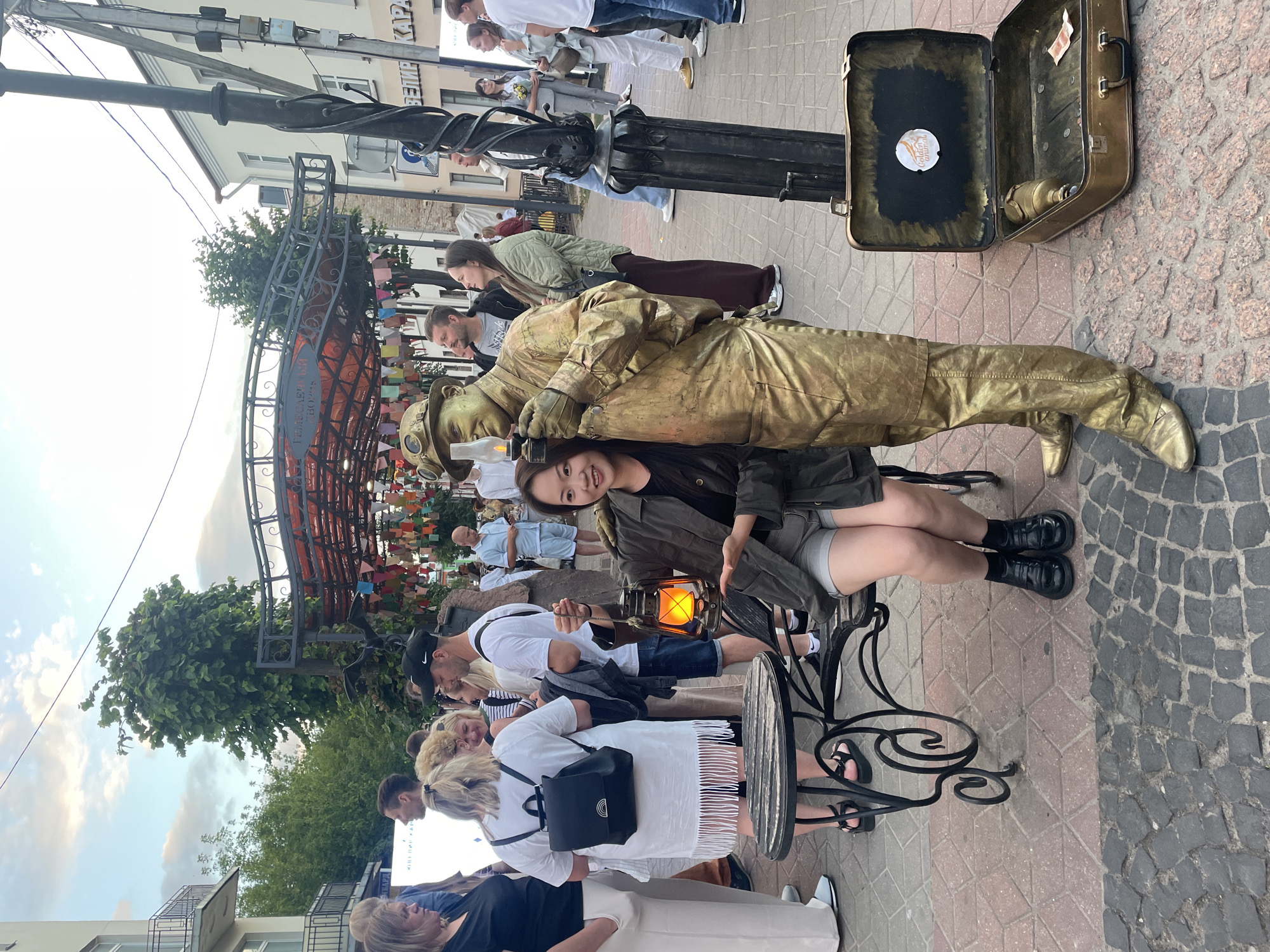 In a city setting, likely Brest, Belarus, a young woman in casual attire sits on a bench beside a street performer dressed as a golden statue. The performer, with goggles and a hat, holds a lit lantern. The woman also holds a lantern, which appears to be the same or similar model as the performer's. They are posing for a photograph, with the woman smiling at the camera. In the background, a crowd of people mills about, suggesting a public event or busy street. An open, vintage suitcase lies on the ground near the performer, possibly for donations. The street is paved with cobblestones. The overall scene suggests a lively street performance in an urban environment.