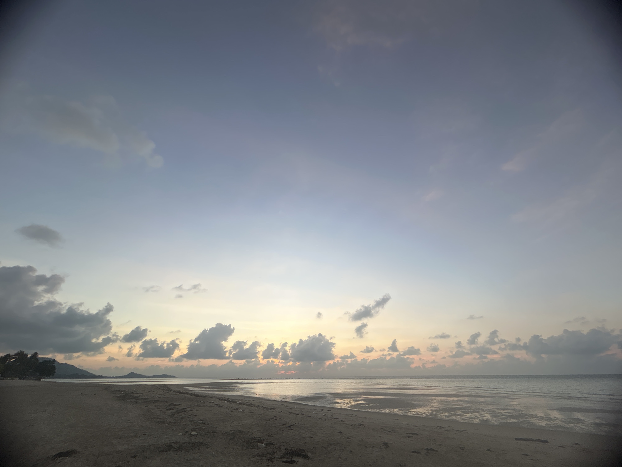 This serene landscape photograph captures a wide, expansive beach scene at either dawn or dusk in Baan Harn, Thailand. The composition is divided into a broad sky, a calm ocean, and a dark sandy foreground.
The sky, occupying the upper two-thirds of the image, displays a beautiful gradient of soft blues, purples, and pale yellows and oranges stretching along the horizon. Scattered cumulus clouds are dispersed across the sky; some appear dark grey, particularly on the left, while those closer to the sun's position are illuminated with gentle hues.
The ocean surface is tranquil, reflecting the sky's colors and appearing almost silvery in the soft light. The tide seems to be low, revealing wide stretches of damp, textured sand with ripples and patterns in the midground and foreground. On the far left of the horizon, a distant landmass with dark silhouettes of trees or dense vegetation leads into low-lying hills or mountains, characteristic of a tropical coastline.
There are no people or specific activities depicted, emphasizing the quiet natural beauty of the moment. The overall atmosphere is one of peace and calm, with the soft, diffused light creating a tranquil ambiance. No visible text is present in the image.