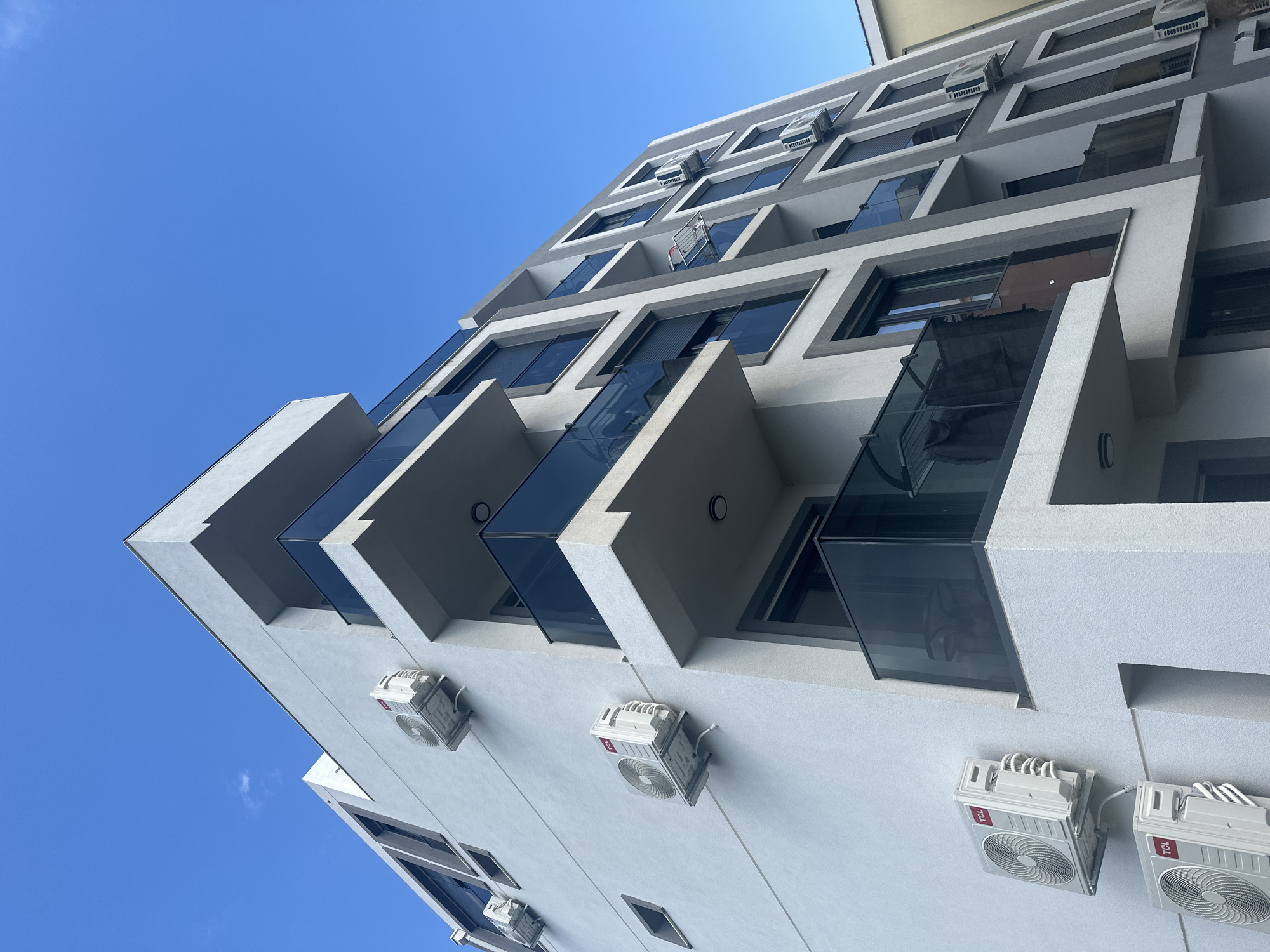 The image depicts a modern building in Bar, Montenegro, characterized by a light gray facade and a series of jutting balconies with glass railings. The building appears to be multi-story, with several windows per floor. Numerous air conditioning units are attached to the exterior walls, beneath the windows and balconies. The sky is a clear, bright blue. A shopping cart can be seen sitting on a balcony.