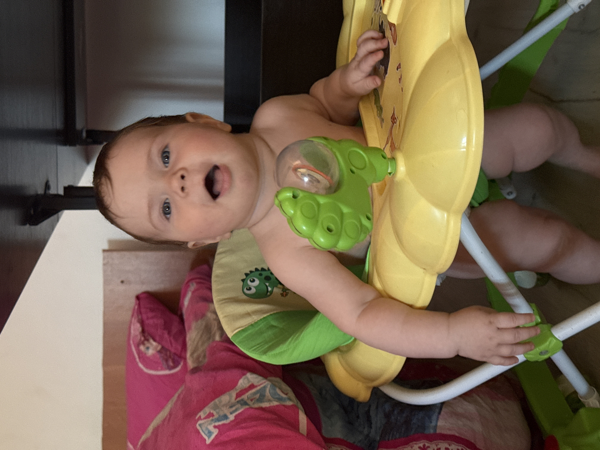 A baby is in a yellow and green baby walker indoors. The baby has fair skin, blue eyes, and an open mouth. The walker has a seat with a dinosaur on it and several toys. The baby's legs are visible below the seat. A bed with a pink and blue patterned comforter is behind the baby. A dark brown piece of furniture is on the left side of the image. There is no information about the location being in Rusalevka, Russia.