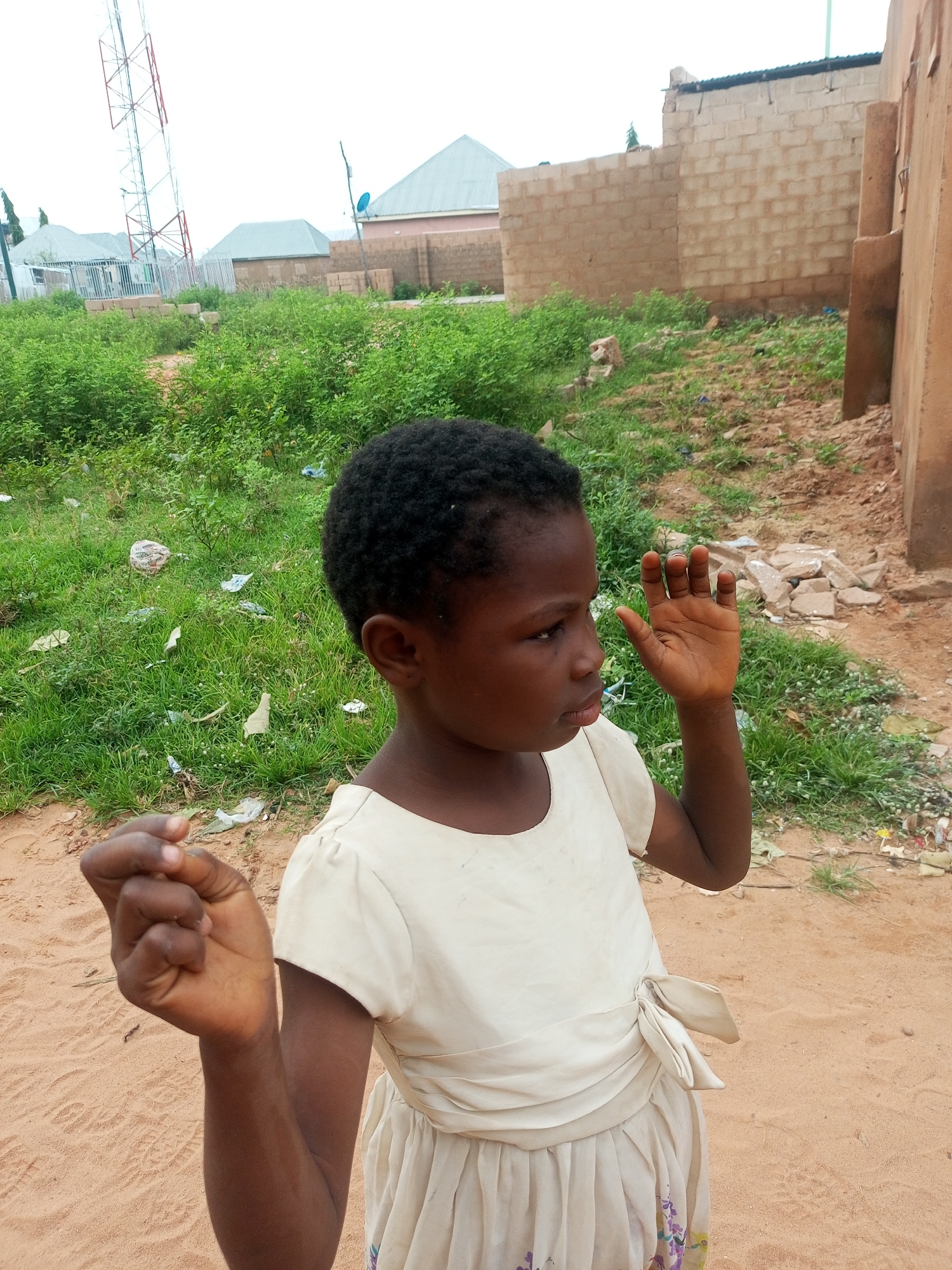 A young girl with dark skin and short, dark hair stands on sandy ground in front of a background of overgrown green vegetation, a brick building, and houses. She is wearing a light-colored, possibly cream or off-white, dress with gathered fabric and a bow at the waist. The girl is looking to her right and has her right hand raised, palm facing forward, as if waving or signaling. Her left hand is clenched into a fist. In the background, a tall telecommunications tower is visible to the left, along with the roofs of several buildings and a partially constructed brick wall. The ground appears to be a mix of sand and dirt, with scattered debris in the vegetation. The sky is overcast. The location is identified as Tempure, Nigeria.