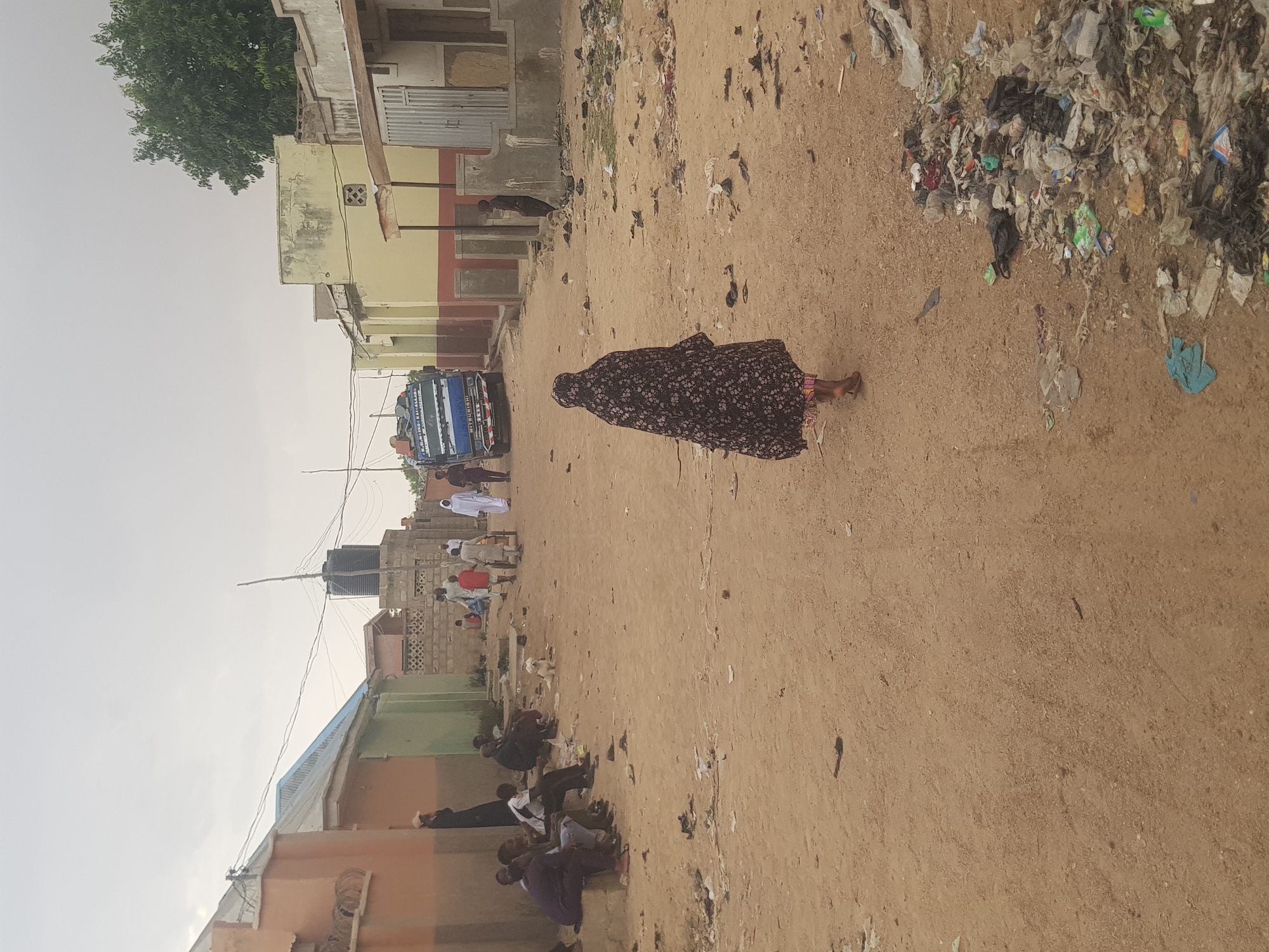 The image depicts a street scene in Dabawa, Nigeria. The unpaved road is the primary surface, covered with scattered litter and debris. A woman in a dark, patterned dress is walking along the road. Further down the road, a group of people are gathered, some sitting on the ground. A blue truck is parked nearby. Buildings line both sides of the street, constructed of cinder blocks and painted in varying colors. The sky is overcast, suggesting daytime. A water tank is visible atop one of the buildings.