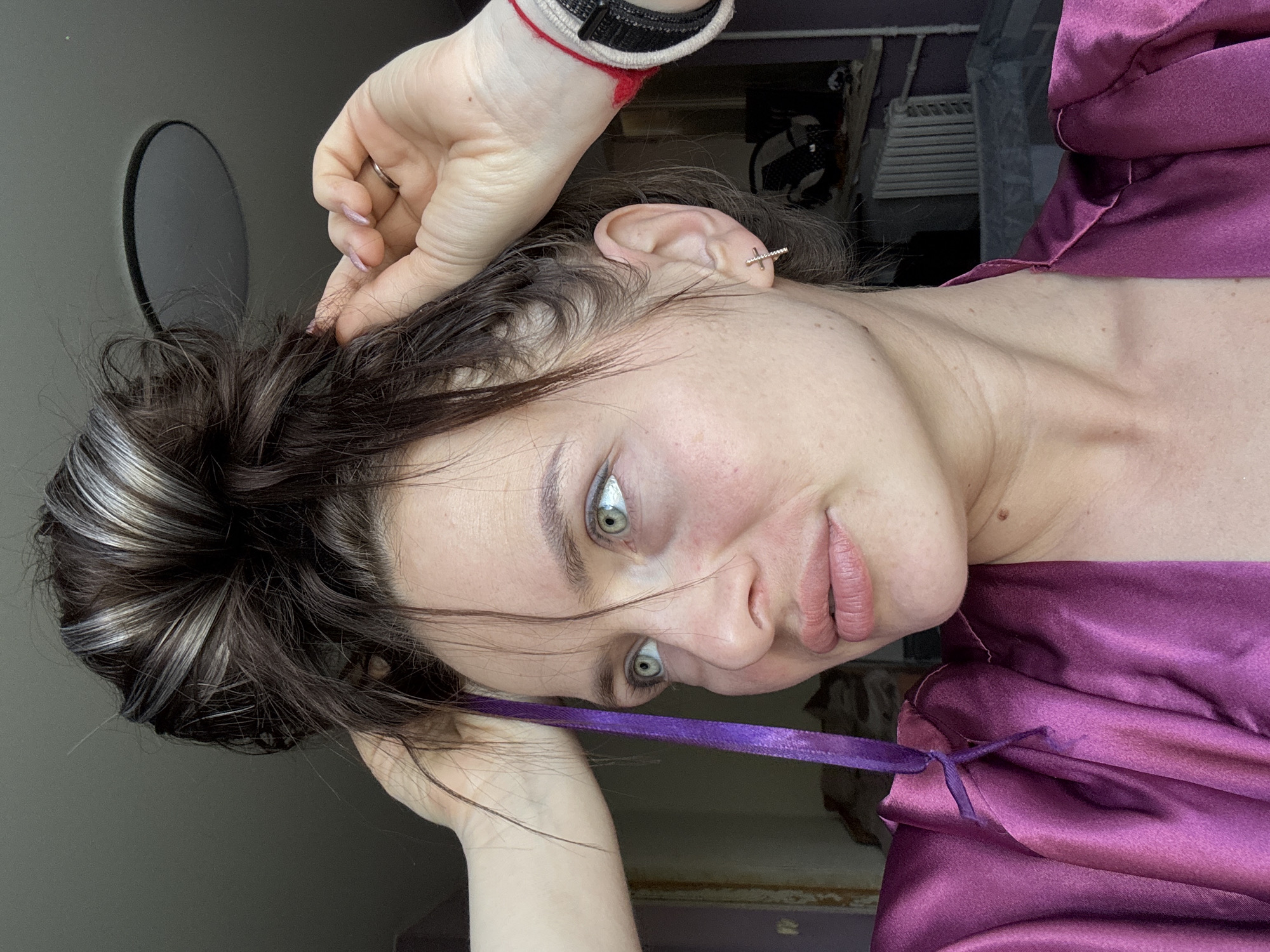 A woman with grey-green eyes, dark hair with silver streaks piled on top of her head, and light skin is seen from a high angle. She is wearing a shiny, purple robe, tied at the chest with a purple ribbon. Her left hand is holding the top of her head, while her right hand is near her ear, and has a black and white bracelet with a red string tied around her wrist. She has a stud earring in her right ear. In the background is a grey wall, a round dark object on the wall, and a glimpse of a room. The location is Rusalevka, Russia.