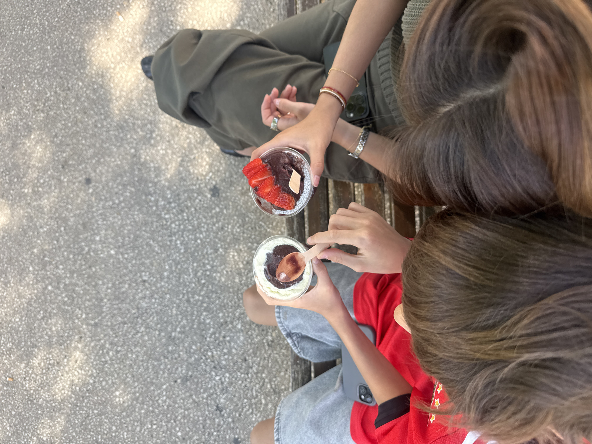 Two individuals sit on a bench. One wears green pants and a knitted top, holding a plastic cup filled with a dark substance topped with chia seeds and sliced strawberries. The other wears a red shirt and grey shorts, holding a cup of layered cream and chocolate with a wooden spoon inserted. The bench is located on a concrete surface with a speckled pattern.