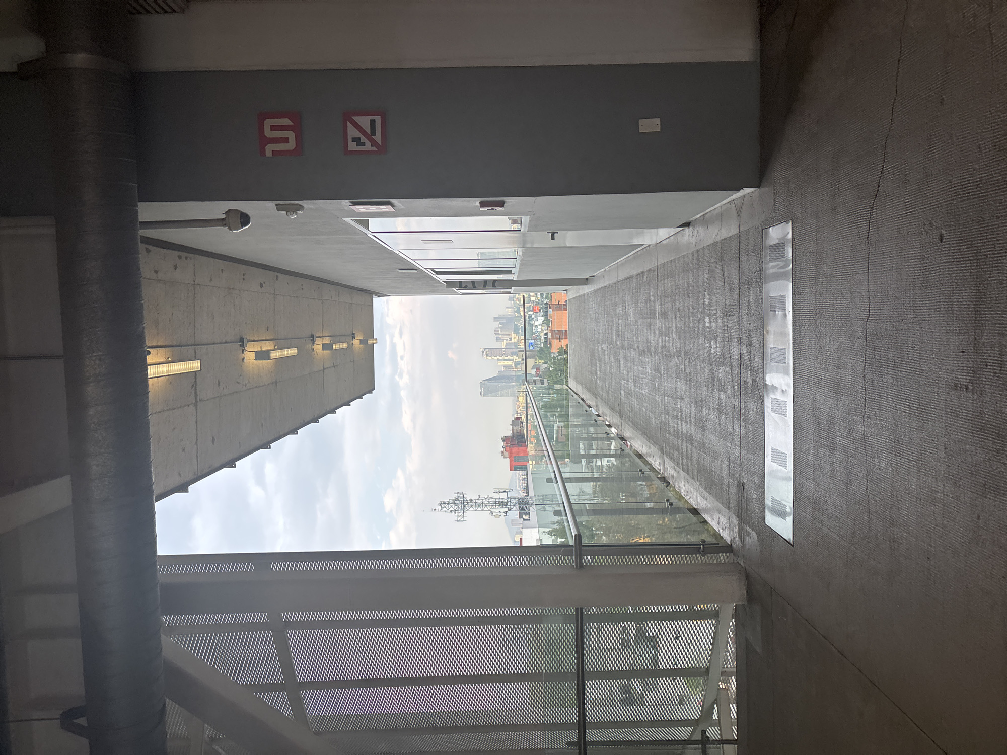 The image depicts a long, narrow concrete passageway leading to an opening revealing a view of Mexico City, Mexico. The passageway has walls on either side; the left wall is concrete with light fixtures, and the right wall is textured concrete. Safety signage is visible on the left wall. A glass barrier lines the edge of the passageway, offering a view of the city's skyline. A metal grate structure is visible in the foreground, partially obscuring the lower portion of the city view. The sky is overcast.