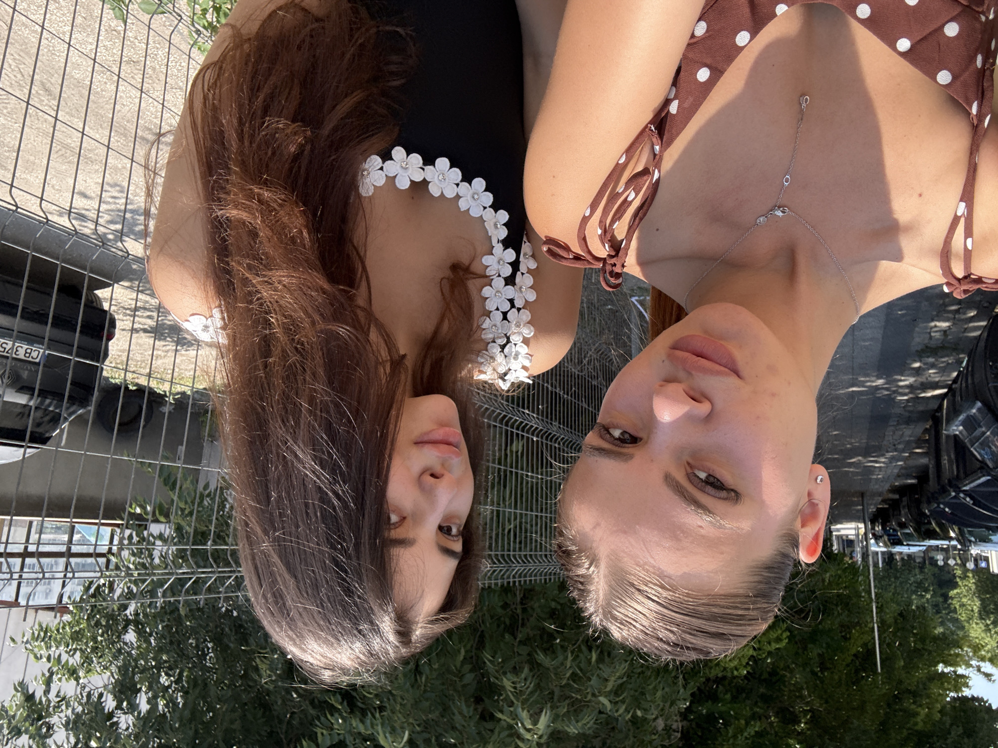 Two young women are posing for a selfie outdoors in a city setting. The woman on the left has her hair pulled back and is wearing a brown polka dot top with tie straps. She is also wearing a silver necklace with a pendant. The woman on the right has long, dark brown hair and is wearing a black top adorned with white flowers and rhinestones. They are standing in front of a metal fence. In the background, there are trees, a sidewalk with trash bins, parked cars, and a building. The license plate on the black car visible in the background reads "CB 375". The presence of specific license plate formatting (CB prefix) suggests the location is likely in Bulgaria, and with the provided context, it is Plovdiv.