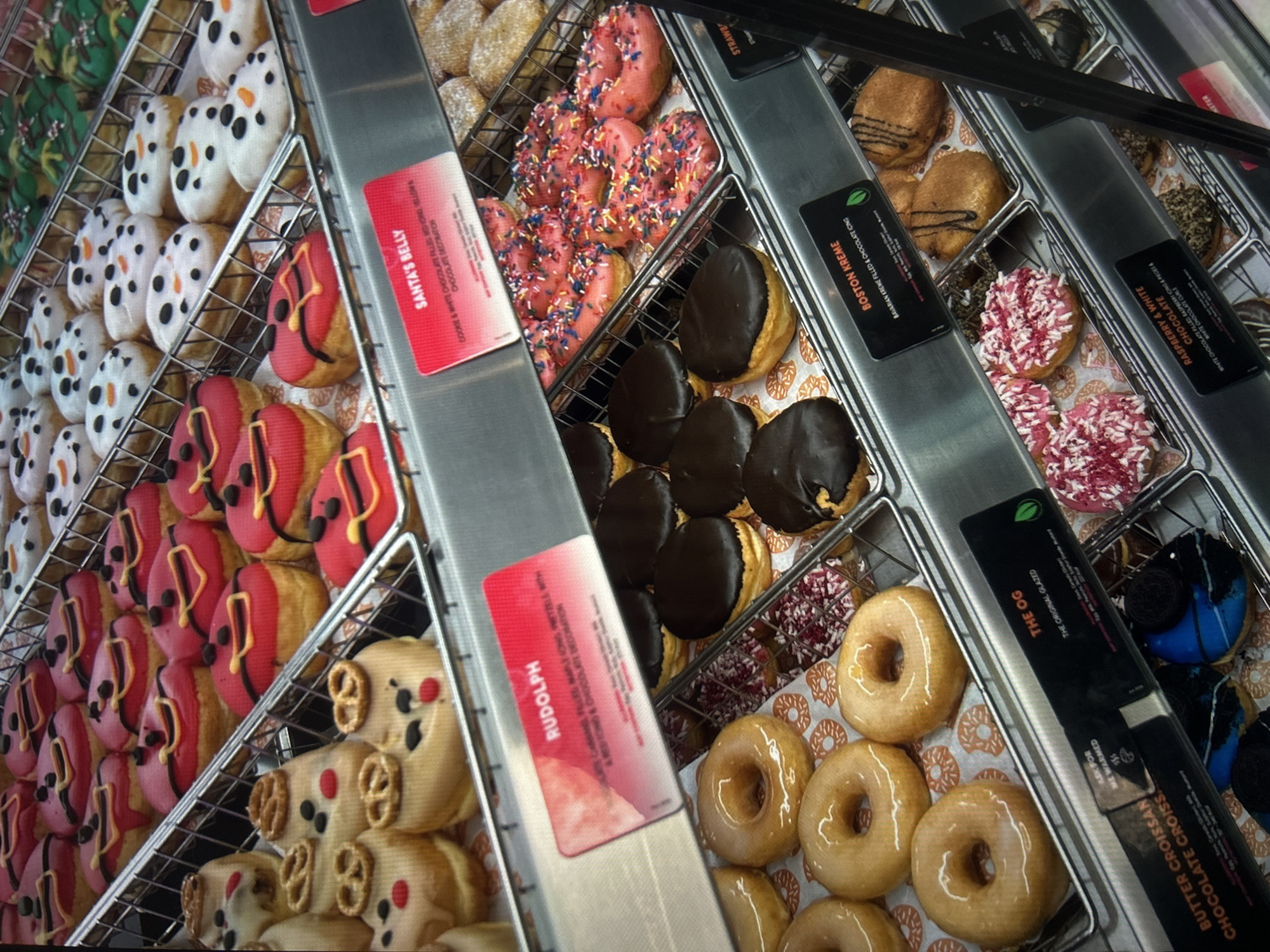The image shows a display case filled with various types of donuts. The donuts are arranged in rows in metal racks. Donuts visible include glazed, red frosted with yellow decoration, snowman-themed, Rudolph-themed, and pink frosted with sprinkles. There are also Boston Kreme donuts with chocolate icing, and raspberry and white frosted donuts with white chocolate curls. Signage indicates "Santa's Belly", "Rudolph", "Boston Kreme", "The OG", and "Raspberry & White" donuts. The labels also include brief descriptions of ingredients. The image does not provide any information to determine a specific location in Gorna Oryahovitsa, Bulgaria.