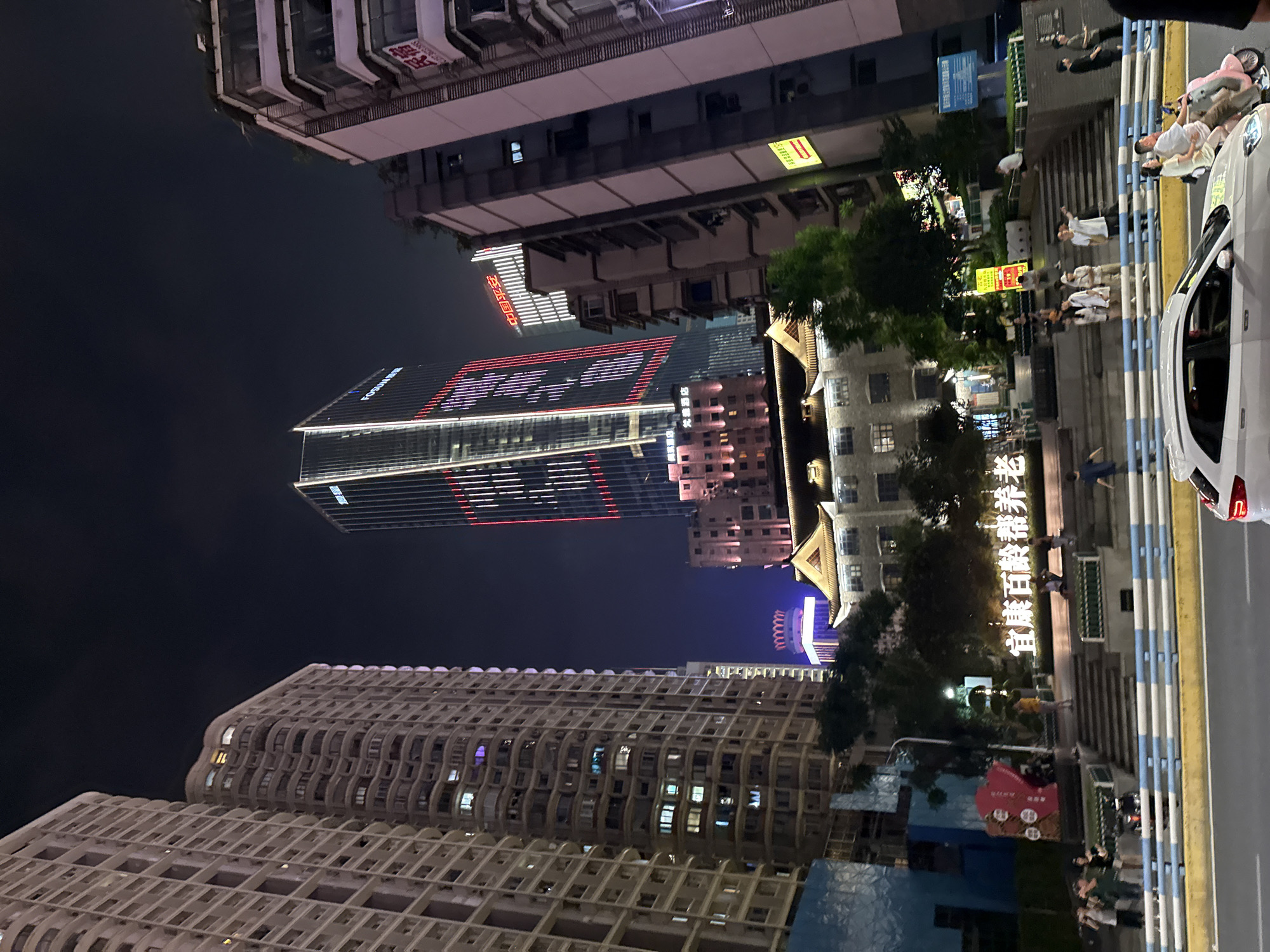 This is a night-time, eye-level, exterior, medium shot of a city street in Yuzhong District, China. The scene is dominated by tall buildings, with a prominent skyscraper displaying red and white lights in the center. To the left, a light-colored, modern apartment building with many windows occupies a significant portion of the frame. To the right, a darker, narrower building stands, with some windows illuminated.
In the foreground, a white car is partially visible, moving from right to left. On the street in front of the buildings, a pink scooter carrying two people is also moving from right to left. There are several people standing and walking on a tiered pedestrian walkway in front of a traditional-style building with a dark, tiled roof. Some of these individuals are interacting, with one person pointing. Behind the walkway, a large sign with white text is illuminated against a dark background. A decorative red structure with circular motifs is visible to the left of the sign. Trees are interspersed throughout the scene. The sky is dark, indicating it is nighttime.