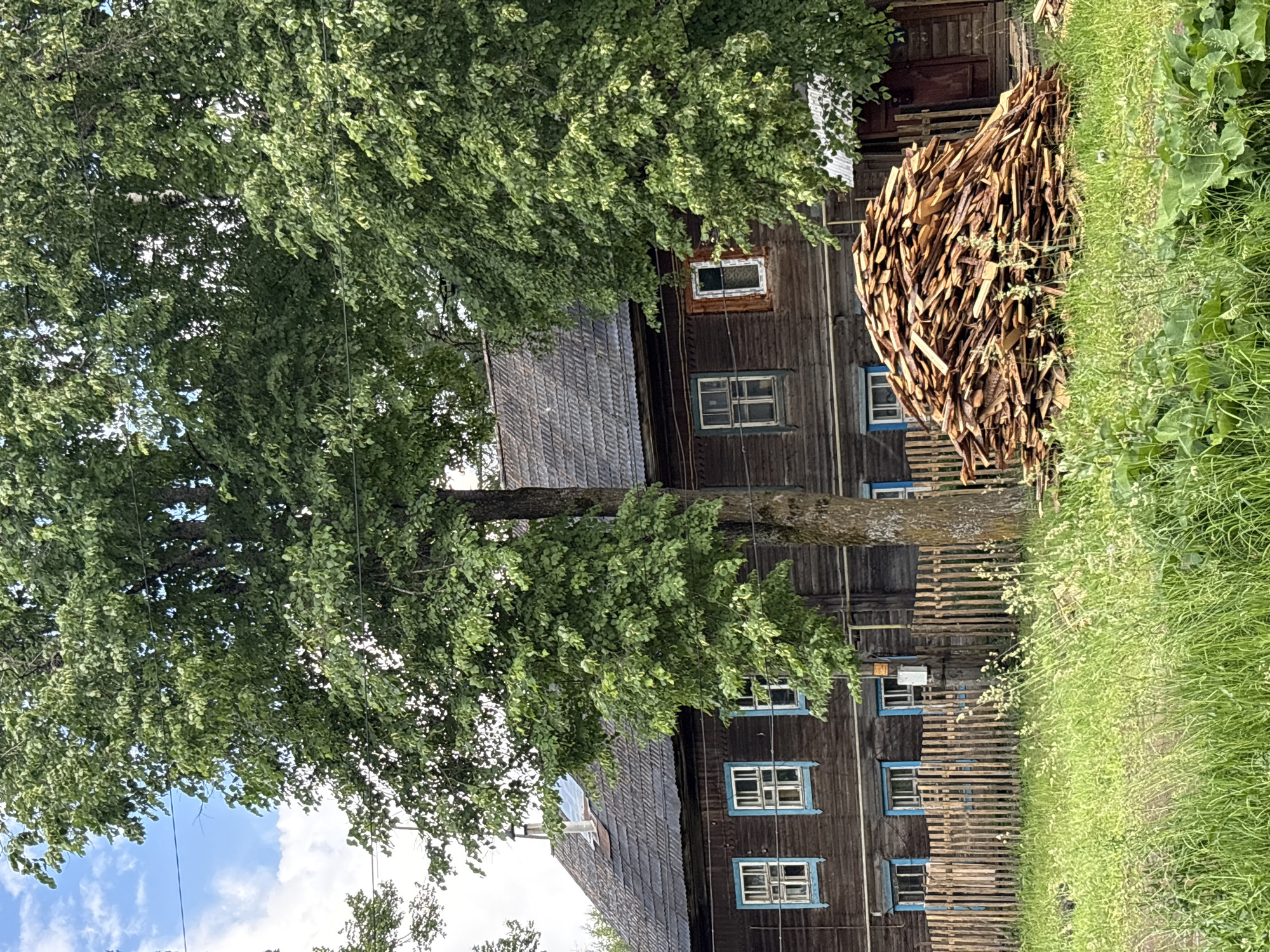 The image shows a wooden house with a dark wooden facade and a grey, shingled roof. There are several windows with blue trim on the front of the house. In front of the house is a wooden fence and a large pile of lumber. Lush green grass and trees surround the scene. The sky above is blue with some white clouds. The photograph appears to be taken in a rural setting.