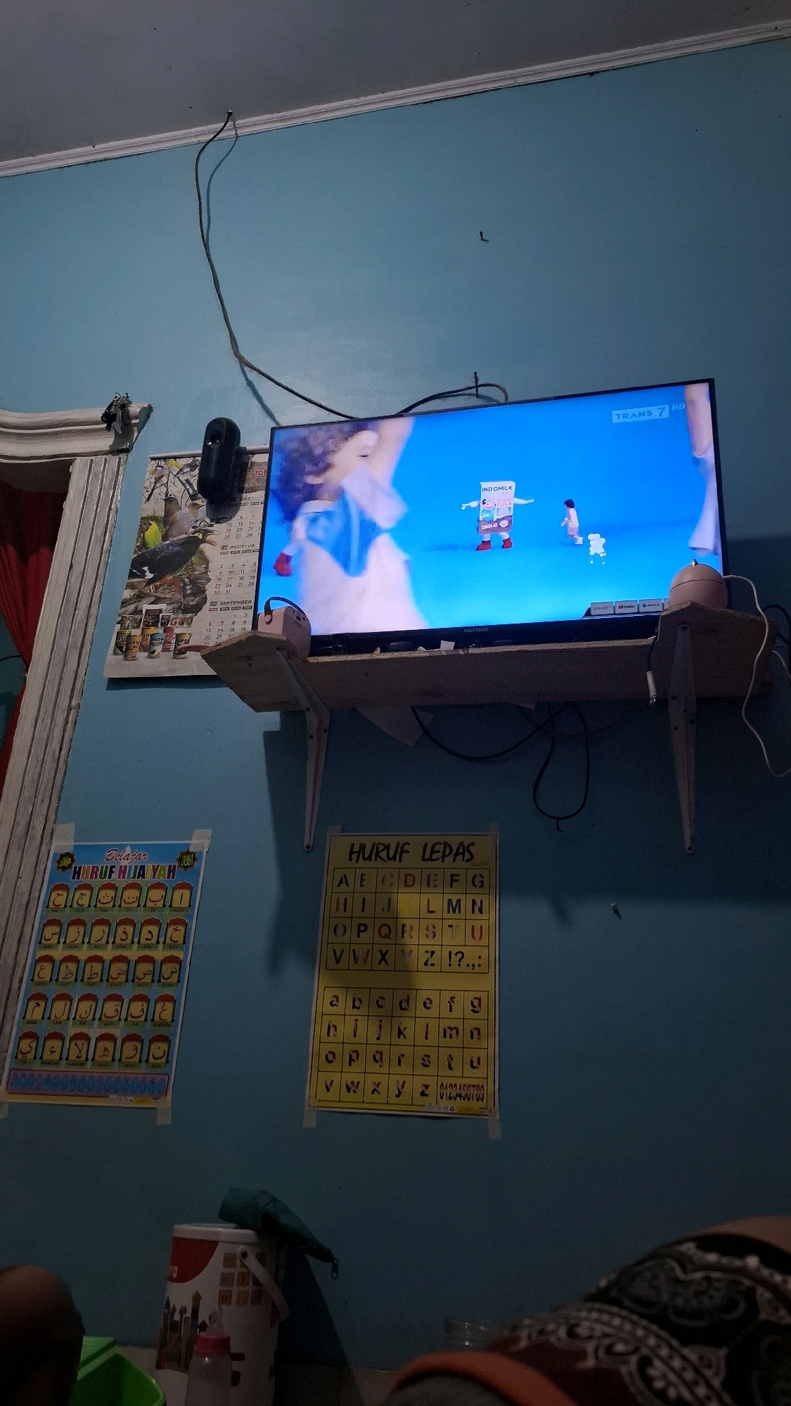 The image depicts an indoor scene in Bandar Lampung, Indonesia, likely a living room. A television set is mounted on the wall and displaying a children's program with animated characters. Below the television, a wooden shelf is installed, holding a pink speaker and some cables. To the left of the television, a calendar is hanging on the wall, featuring a picture of a bird. Two educational posters are also visible: one on the left with Arabic alphabets and another on the right showing English alphabets. In the foreground, a person's legs and shoulder are visible, suggesting they are sitting or lying down. Various objects like buckets and bottles are scattered around the floor. The room is painted a light blue color.