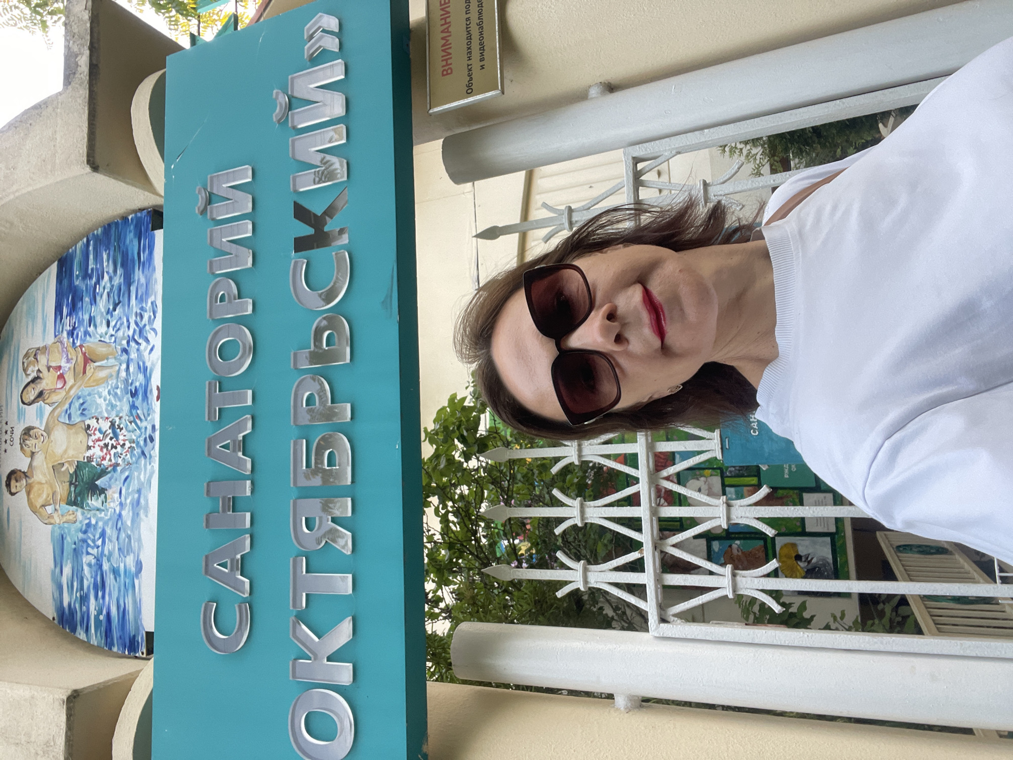 The image shows a woman in front of a sign that reads "САНАТОРИЙ ОКТЯБРЬСКИЙ" which translates to "Sanatorium Oktyabrsky". This sanatorium is located in the city of Sochi, Russia. The sign is on a teal background and the letters are silver and reflective. Above the sign is a mural depicting people in the water. The woman is wearing sunglasses and a white shirt. Behind her is a white metal fence and trees. To the right of the sign, there is a warning sign in Russian that reads "ВНИМАНИЕ. Объект находится под видеонаблюдением и записью." which translates to "ATTENTION. The facility is under video surveillance and recording."