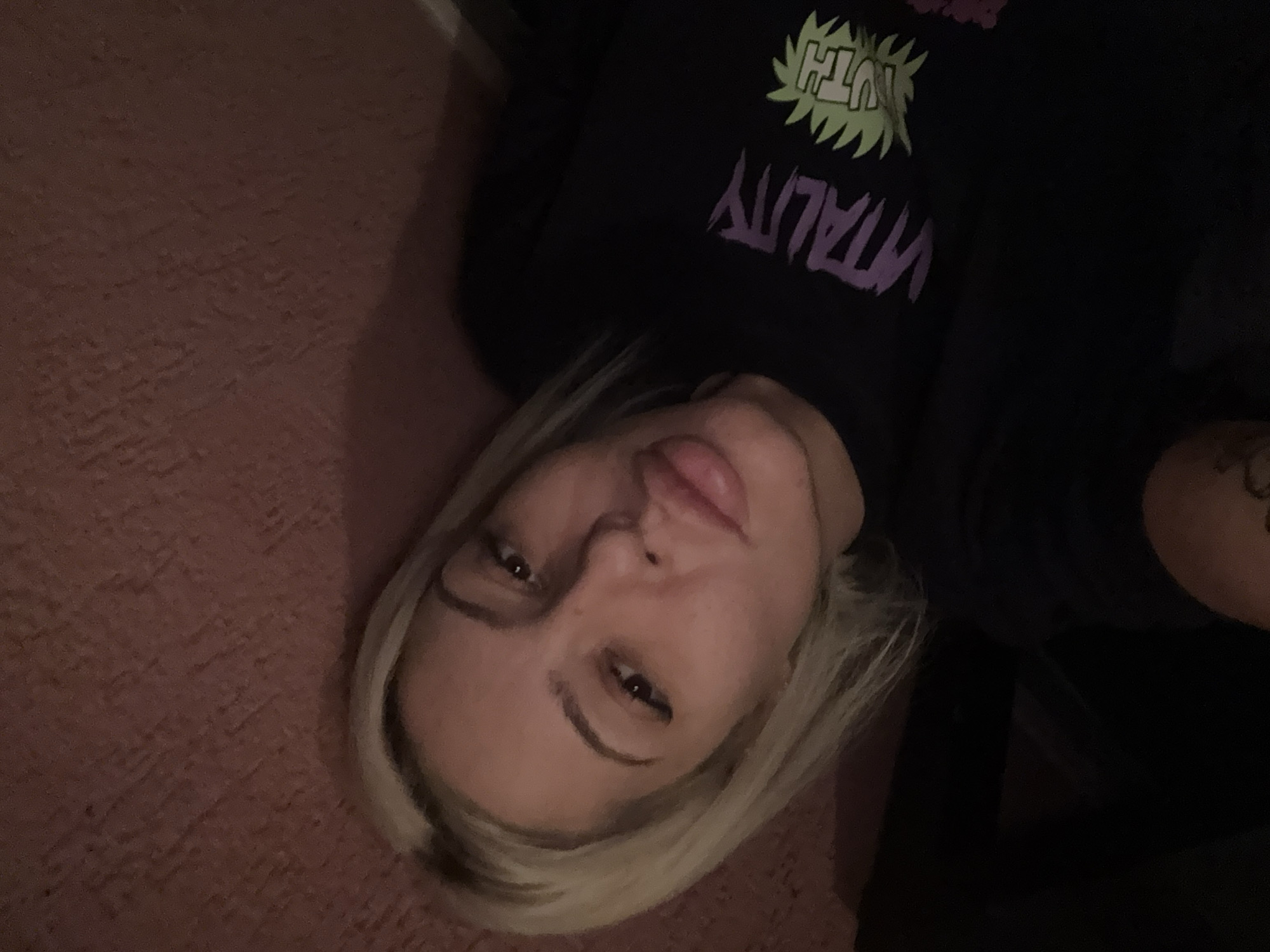 A woman with blonde hair is lying on a pink carpet. She is wearing a black sweatshirt with the words "VITALITY" and "YOUTH" printed on it. A tattoo is visible on her right arm. The location in the city of Plovdiv, Bulgaria, cannot be determined from the image.