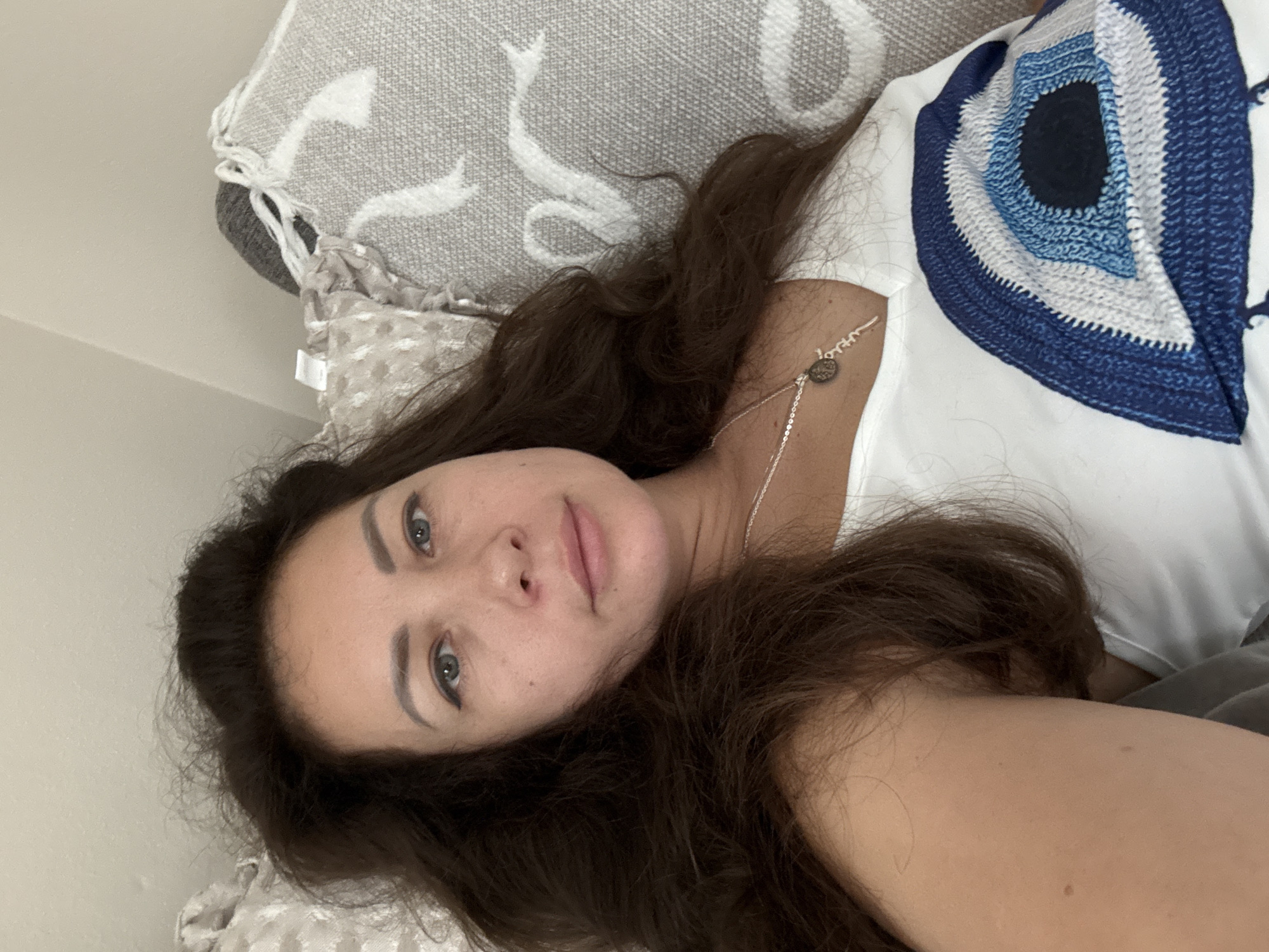 The media file is a selfie taken by a woman lying on a couch. The woman has long, dark brown hair and is wearing a white t-shirt with a blue and white evil eye design. She is looking at the camera with a neutral expression. The background shows a patterned pillow and a wall. The lighting suggests it is daytime, and the indoor setting implies a relaxed, at-home environment. There is no visible text in the image. The woman is the sole subject of the media file, and the activity is a casual pose for a photograph.