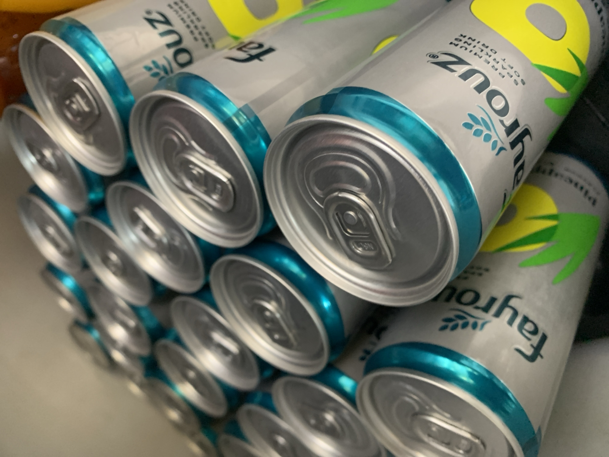 A collection of aluminum cans with silver tops and teal blue accents are arranged in a staggered pattern. The cans have "Fayrouz" printed on them in dark blue lettering, along with smaller text that includes "PREMIUM SPARKLING SOFT DRINK." Some cans also feature yellow and green graphic elements that appear to be fruit-related. The cans are tightly packed together, suggesting they are stored or displayed in a confined space. The lighting is somewhat dim, with some highlights reflecting off the metallic surfaces of the cans. The background is indistinct and appears to be a neutral, light-colored surface. There are no people visible in the image. The scene is likely indoors, possibly in a refrigerator, pantry, or store display.