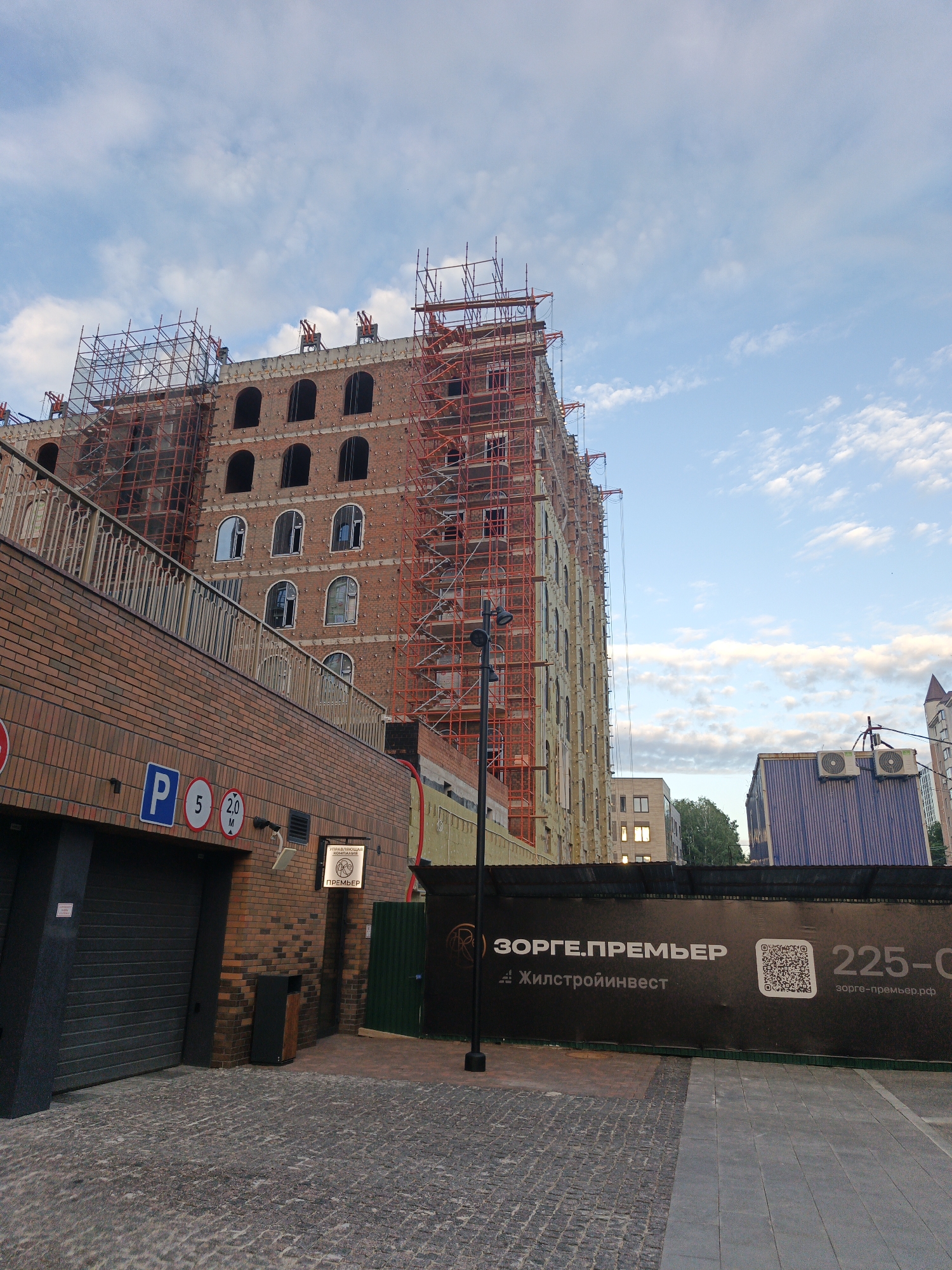 The image depicts an unfinished multi-story building, likely a residential complex, under construction in Ufa, Russia. The building is clad in red brick and is surrounded by scaffolding. Several arched windows are visible, some of which are boarded up, while others appear to be open. The ground level features a brick facade with a dark garage door and a pedestrian entrance. Parking signs, including a "P" symbol and height restrictions, are posted near the entrance. A large black banner with white text and a QR code is displayed on a fence in the foreground, advertising the construction company "ЗОРГЕ.ПРЕМЬЕР" and its website "zorge-premier.рф". The sky is partly cloudy, suggesting it is daytime. Several other buildings, including one with blue siding and air conditioning units, are visible in the background. The overall scene suggests an active construction site in an urban environment.