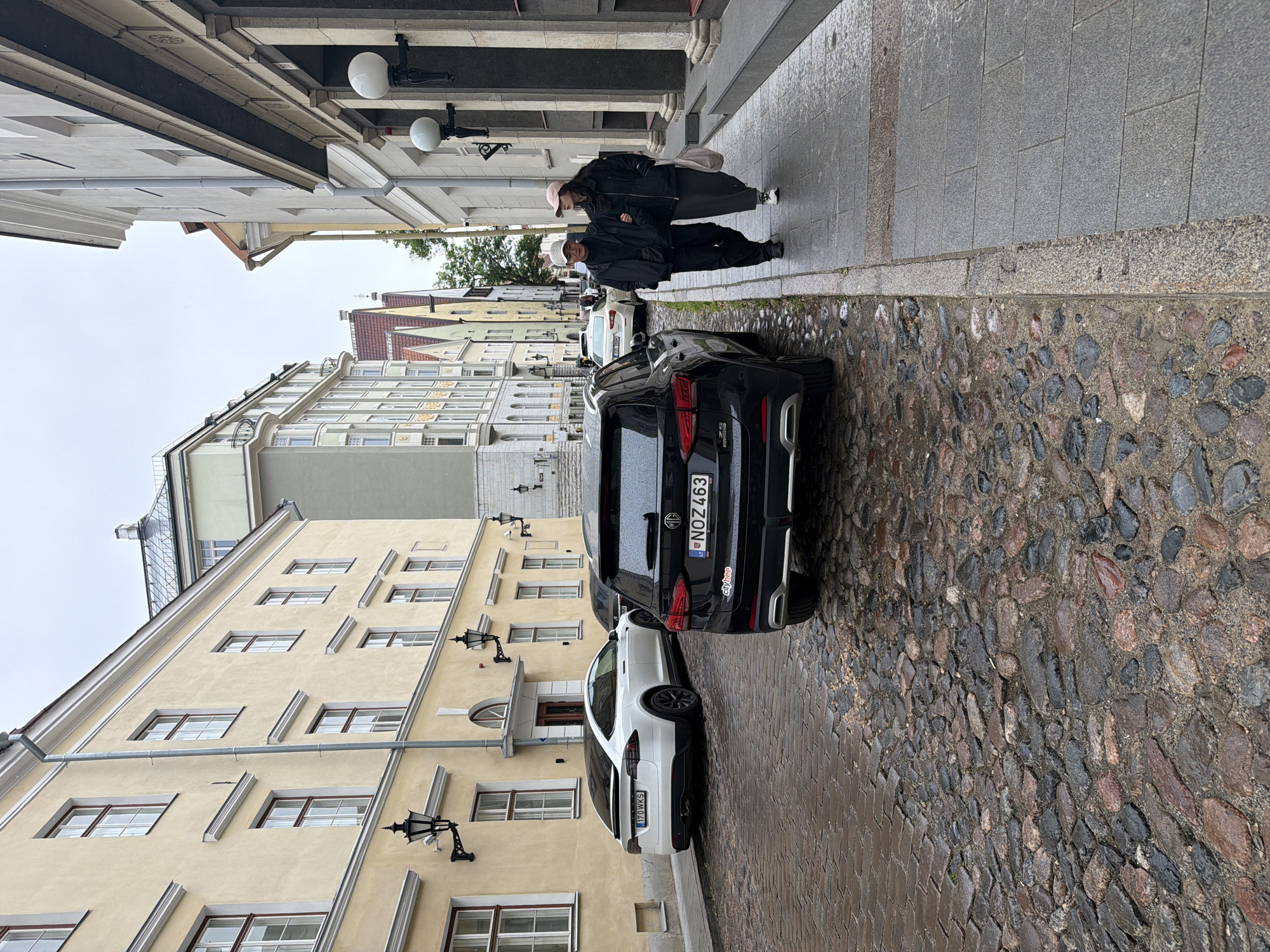 This is a full, eye-level, outdoor, daytime shot of a cobblestone street in Tallinn, Estonia. The street is wet, suggesting it has recently rained. On the left side of the street, there are parked cars, including a white Tesla and a black SUV. Beyond the cars is a row of historic buildings with yellow facades. On the right side of the street is a sidewalk with two people, a man and a woman, walking side-by-side away from the camera. The man is wearing a black jacket, black pants, and a light-colored baseball cap. The woman is wearing a black jacket, a long gray skirt, and a light-colored baseball cap. She is carrying a tote bag over her shoulder. The buildings on the right are also historic, with ornate architectural details. The sky is overcast. The image appears to have been taken from the perspective of someone standing on the sidewalk, looking down the street.
