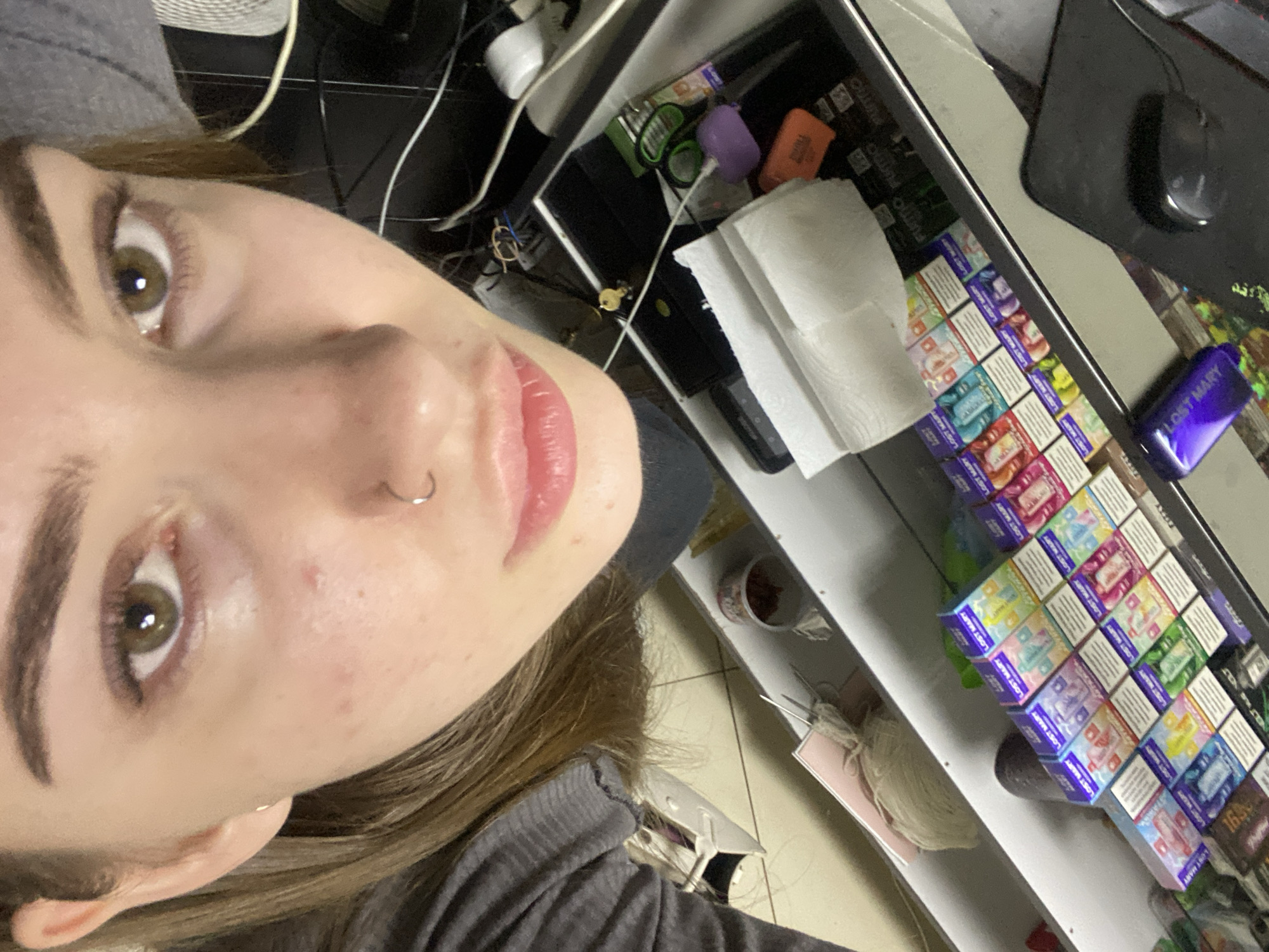 The image is a close-up, overhead view of a young woman's face and upper torso, positioned above a display of vaping products. The woman has light skin, brown eyes, and light brown hair. She is wearing a gray top and has a nose ring. The background consists of a desk or counter with various electronic devices and cables, along with the aforementioned vaping products, which are predominantly small, colorful boxes. The setting appears to be an indoor retail environment, possibly a convenience store or a smoke shop, and the product packaging suggests it could be located in Дьяково, Russia, though this cannot be confirmed solely from the visual information. The image is taken from a high angle, looking down at the woman and the merchandise.