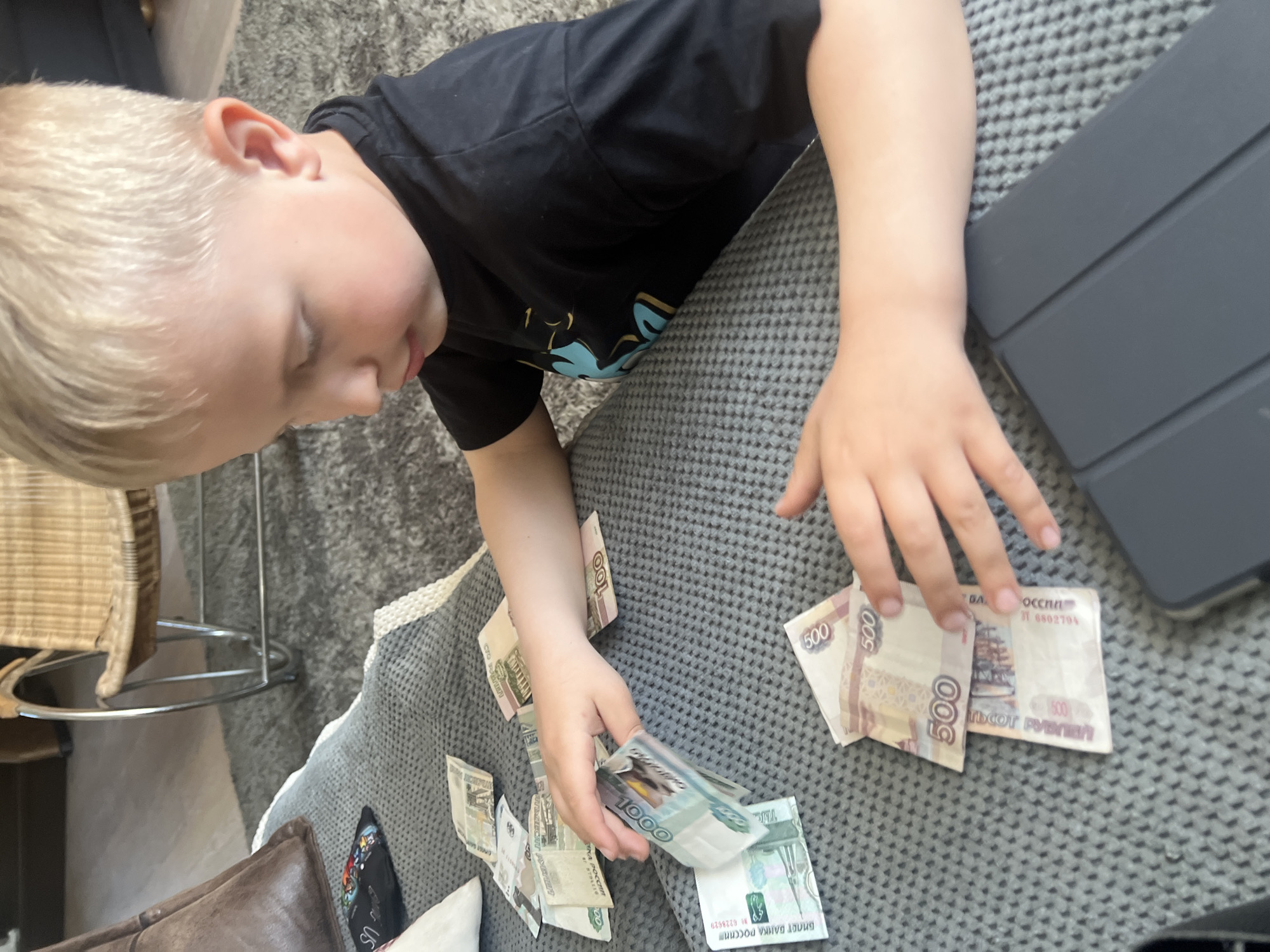 A child is on a grey patterned surface, possibly a couch or rug, with several Russian Ruble bills scattered around. The denominations visible include 500 and 1000 Rubles. The child is wearing a dark t-shirt with a design and has blond hair. The location is undetermined, and there are other objects in the periphery, such as a tablet, part of a chair, and a leather object. The child is interacting with the money, likely counting or playing with it.