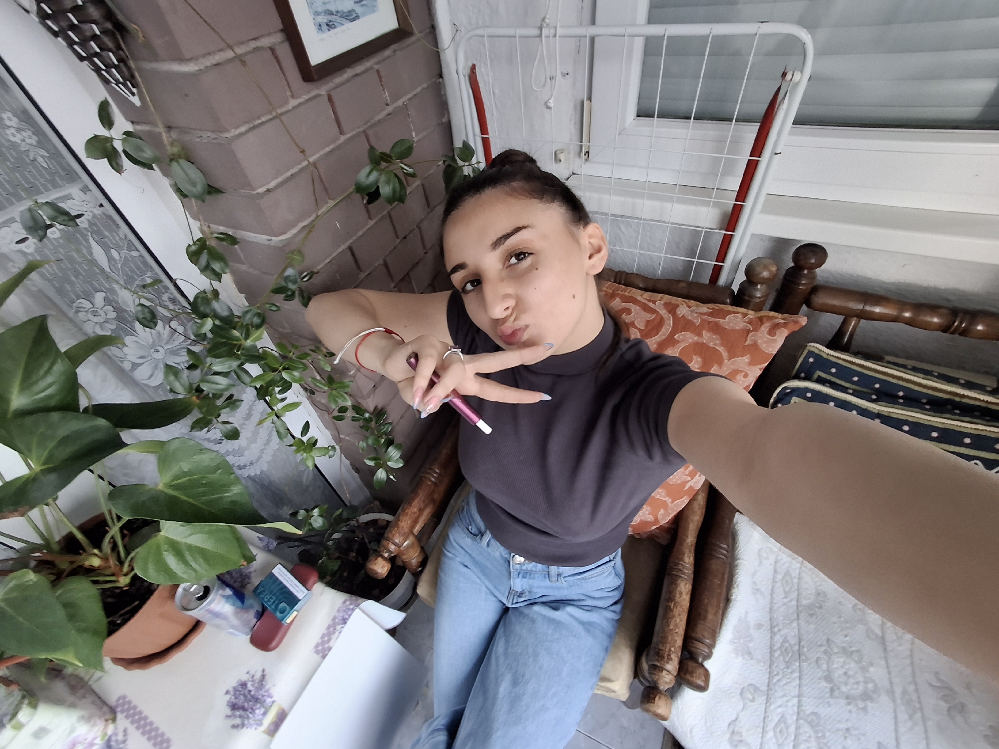 A young woman is taking a selfie. She has her hair in a bun and is wearing a dark-colored top and light blue jeans. She is making a peace sign with her left hand and holding a pen in the same hand. She is puckering her lips for the camera. She is sitting in a wooden chair with patterned cushions. Behind her is a drying rack next to a window. To the left is a brick wall with some greenery growing on it. There is also a potted plant on a table next to her. Based on the architectural style and plant selection, the location is likely an outdoor balcony in an urban area. Without further information, it is impossible to confirm that this photo was taken in Sofia, Bulgaria, and pinpointing the exact location is impossible.