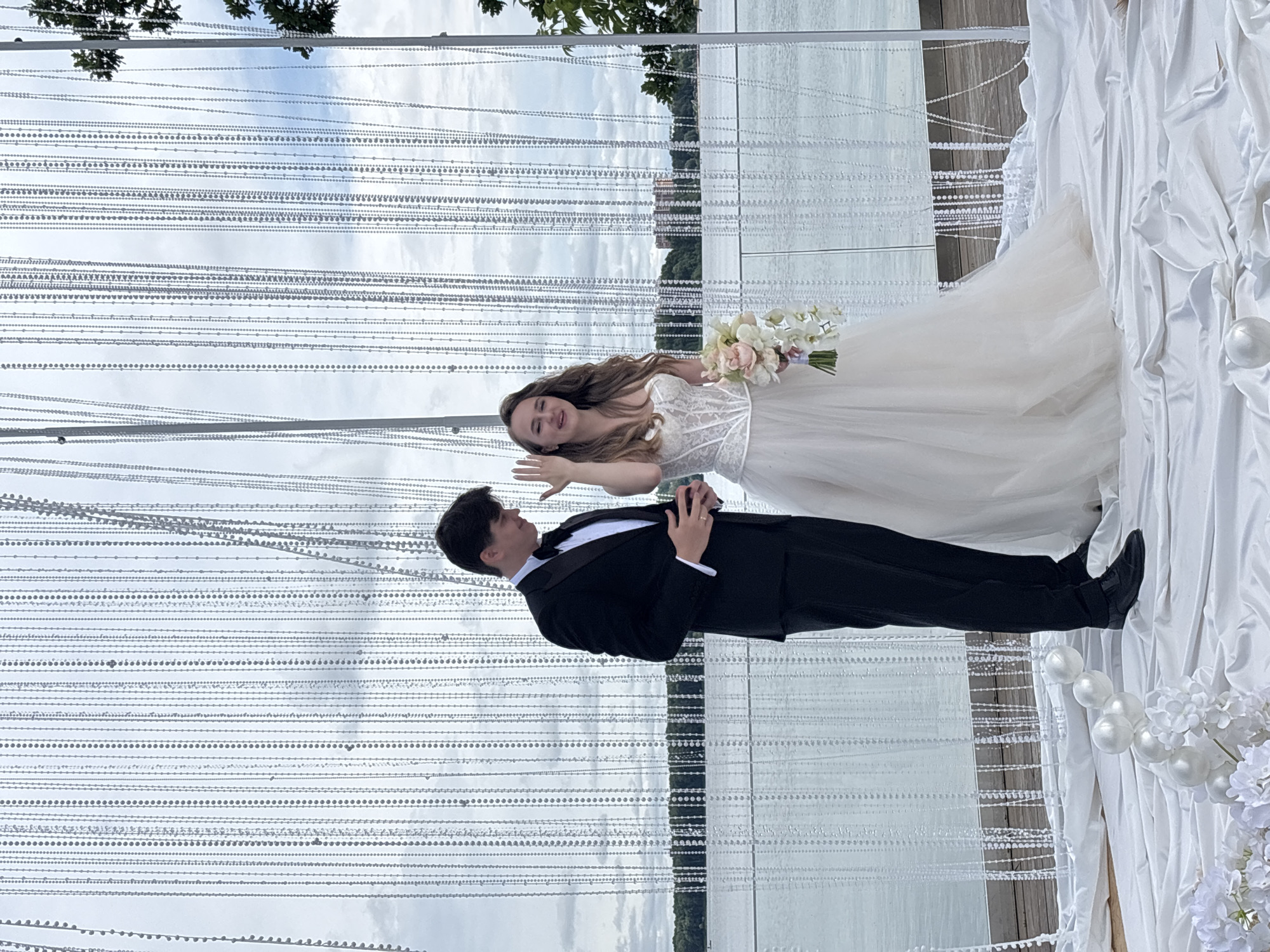 In Osipovo, Russia, a wedding ceremony is taking place outdoors by a body of water. A man in a black tuxedo and bow tie stands to the left, looking down at his hands. To his right, a woman in a white wedding dress holds a bouquet of flowers and raises her left hand, showing her ring finger. They are positioned in front of a backdrop of cascading clear beads, with a hazy sky and trees visible in the distance beyond the water. The foreground is draped with white fabric, adorned with white floral arrangements and spherical decorations.