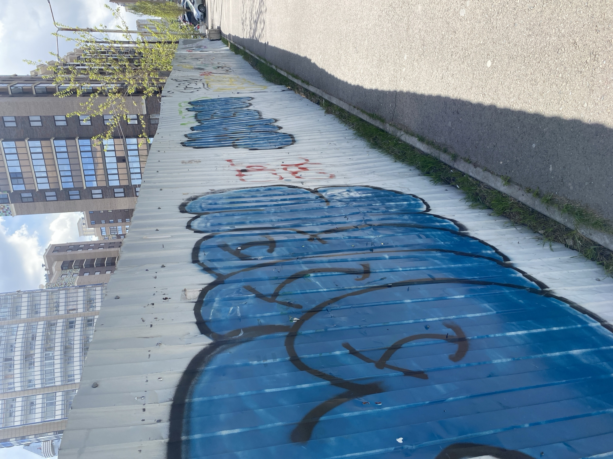 The image depicts a corrugated metal fence with blue graffiti running along its length, positioned alongside a sidewalk and street. Tall buildings, including a high-rise apartment complex, are visible in the background, suggesting an urban setting. Based on the architectural style of the buildings, this scene is likely located in Krasnoyarsk, Russia.
