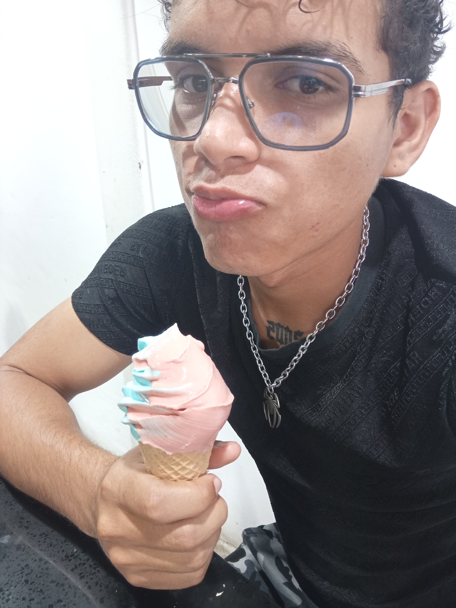 An indoor, close-up portrait shows a young man holding an ice cream cone, looking directly at the camera with a playful, pouting expression. He has light skin, dark curly hair, and wears square-framed glasses with clear lenses. His outfit includes a black short-sleeved shirt with a subtle textured pattern and a silver chain necklace featuring a spider-like pendant. A tattoo, appearing as stylized text or numbers (not clearly legible), is visible on his neck.
In his right hand, the man holds an ice cream cone filled with soft-serve ice cream, which is distinctly colored in pastel pink and light blue. The background is minimalist, featuring a plain white wall behind him and a dark, possibly grey, surface in the lower foreground. The lighting is bright and even, indicating an indoor setting. The image captures a casual moment, potentially in a home or a simple shop in Juan Griego, Venezuela.