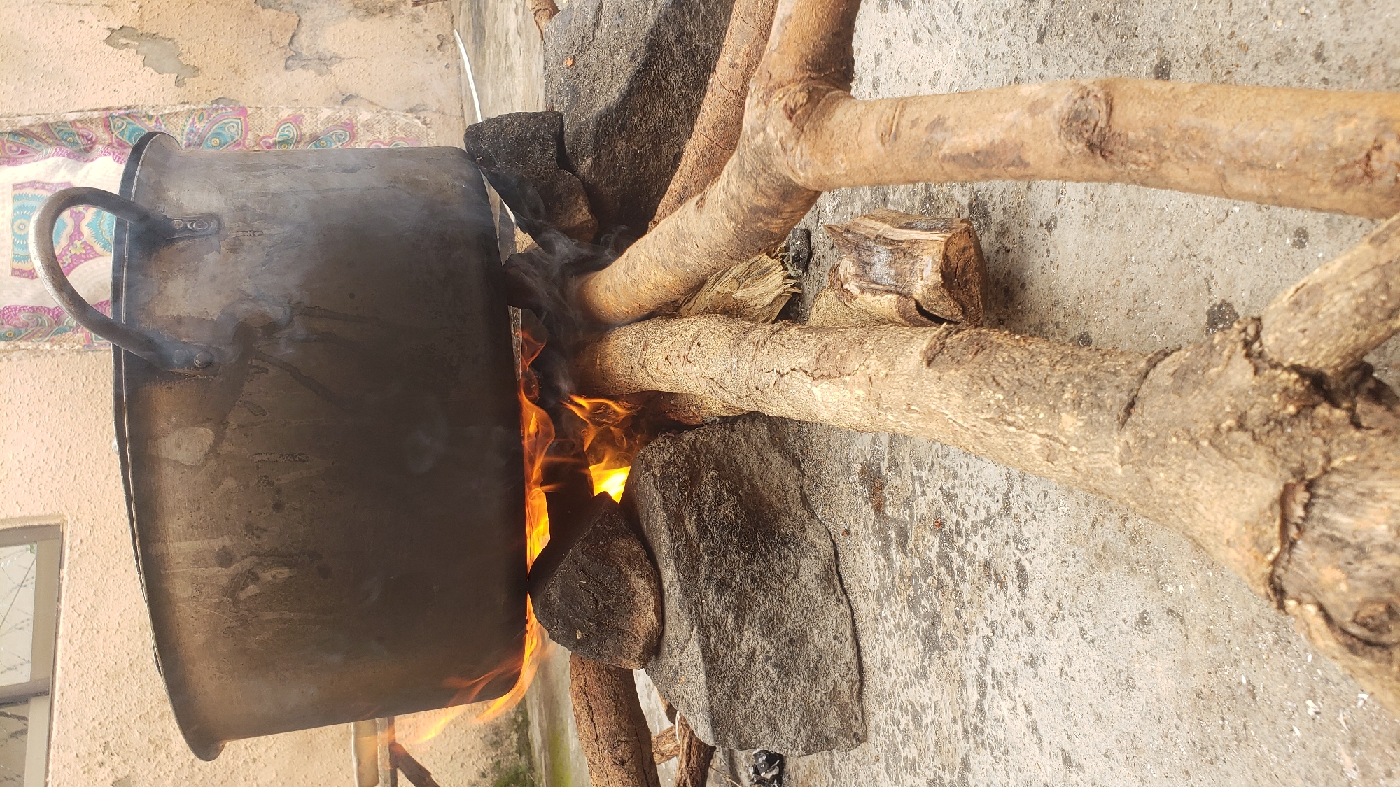 In Sabon Tasha, Nigeria, an outdoor cooking scene unfolds on a concrete surface. A large, dark, metallic pot, showing signs of wear and soot, is suspended over an open fire by rough-hewn stones. The fire beneath glows orange and yellow, with wisps of smoke rising around the pot. Several thick, dry branches are arranged to support the stones and form the base of the makeshift stove. In the background, a textured wall with a window is visible, alongside a colorful piece of fabric hanging, possibly a tapestry or curtain. The overall impression is one of resourcefulness and traditional cooking methods.