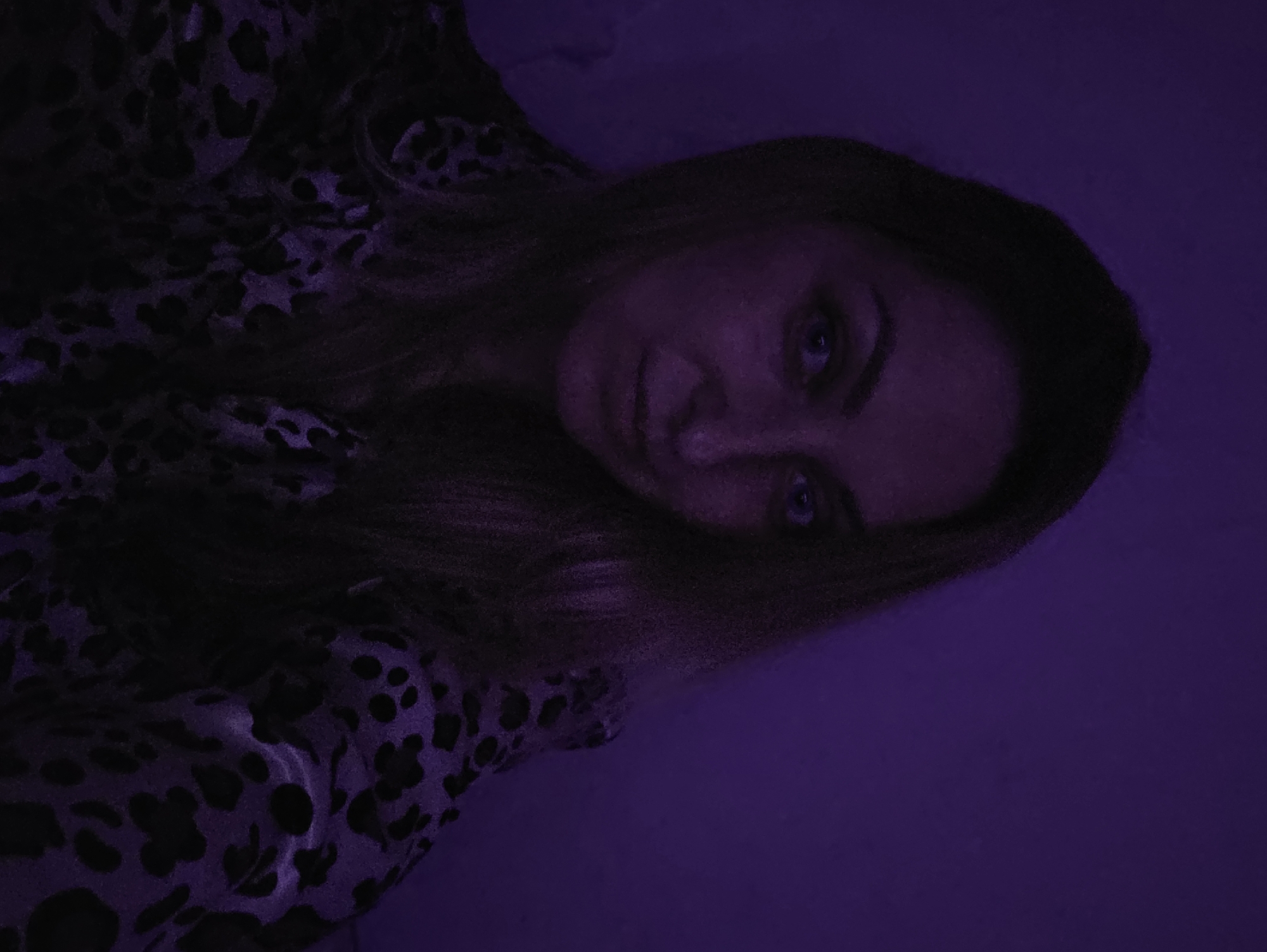 The scene is situated in the city of Minsk, Belarus. A single adult female is depicted in a medium close-up, facing the viewer with a neutral expression. She has long, dark blonde to light brown hair, parted on the right and falling over her shoulders, partially obscuring the right side of her chest. Her eyes, appearing blue or green under the dominant lighting, are directed straight into the camera. She is wearing a dark garment featuring an animal print pattern, specifically black spots on a dark purple or black background, consistent with a leopard or cheetah print. The fabric appears to be satin or a similar material, with subtle sheen. The environment is indoors, characterized by very low light conditions and a uniform, strong purple illumination emanating from an unseen source, bathing the entire scene in deep violet hues. The background is a plain wall, rendered entirely in purple due to the lighting, with no discernible objects or features. The left side of her face from the viewer's perspective is slightly more shadowed.