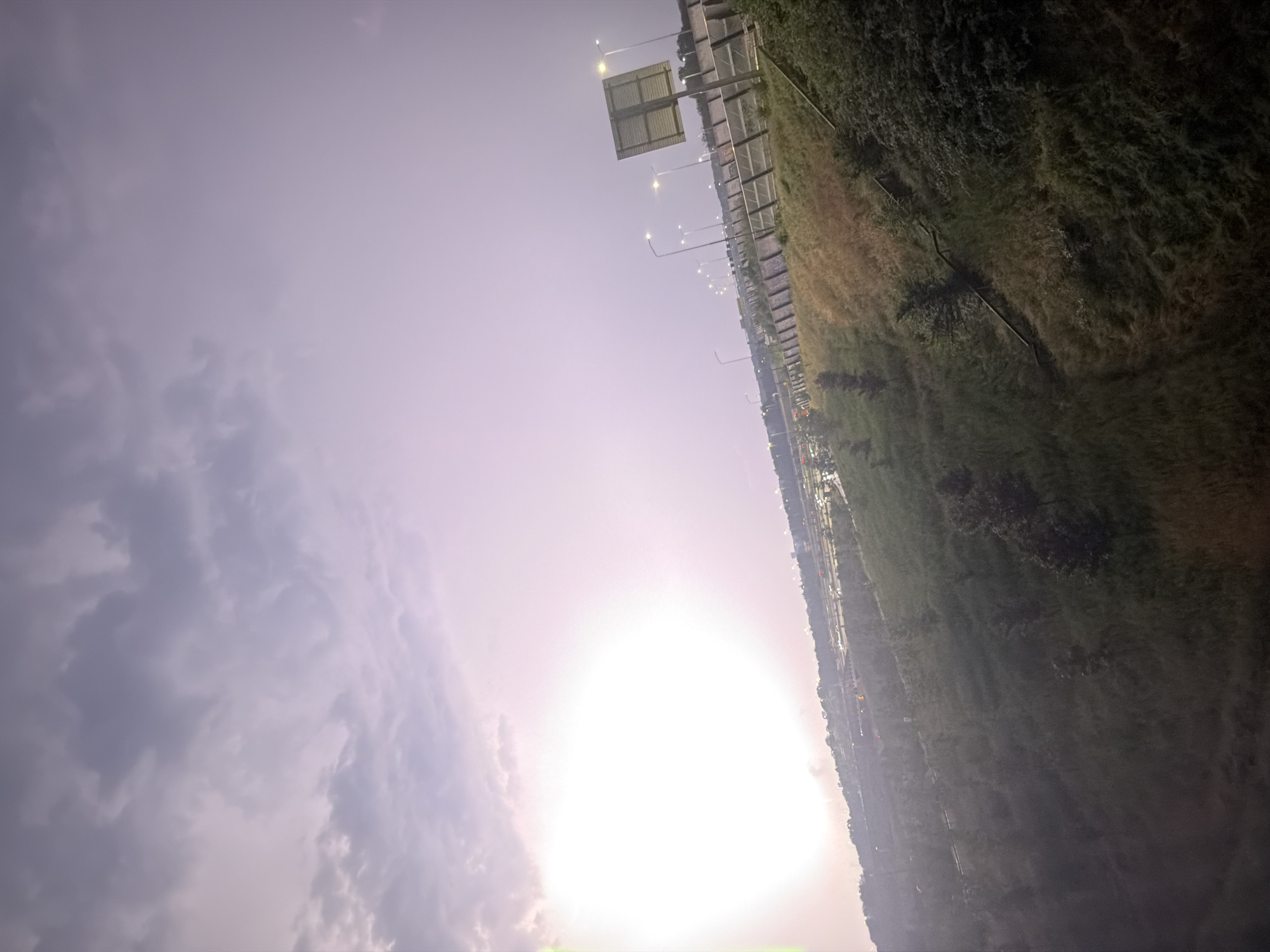 The image captures a scene from Badhoevedorp, Netherlands, likely near a highway or major road. The perspective is from an elevated position looking down and across a sloping grassy embankment. The sky dominates the upper portion of the image, displaying a gradient of soft purple and pink hues, indicative of either dawn or dusk. Large, diffused clouds are present.
In the mid-ground, a highway or roadway is visible, lined with streetlights that are illuminated. Several vehicles are present on the road, their lights suggesting some traffic. A tall signpost with a rectangular sign is positioned on the right side of the road. The embankment is covered with a mix of green and dry grass, with scattered trees and bushes. A set of stairs or a pathway is visible winding down the embankment on the right side of the image.
In the distance, the urban landscape of Badhoevedorp is suggested by buildings and more distant lights, though details are obscured by haze or distance. The overall mood is serene, with the soft lighting and natural elements contrasting with the man-made infrastructure. There are no people visible in the frame. The environment appears to be an edge of a city, where nature meets urban development.