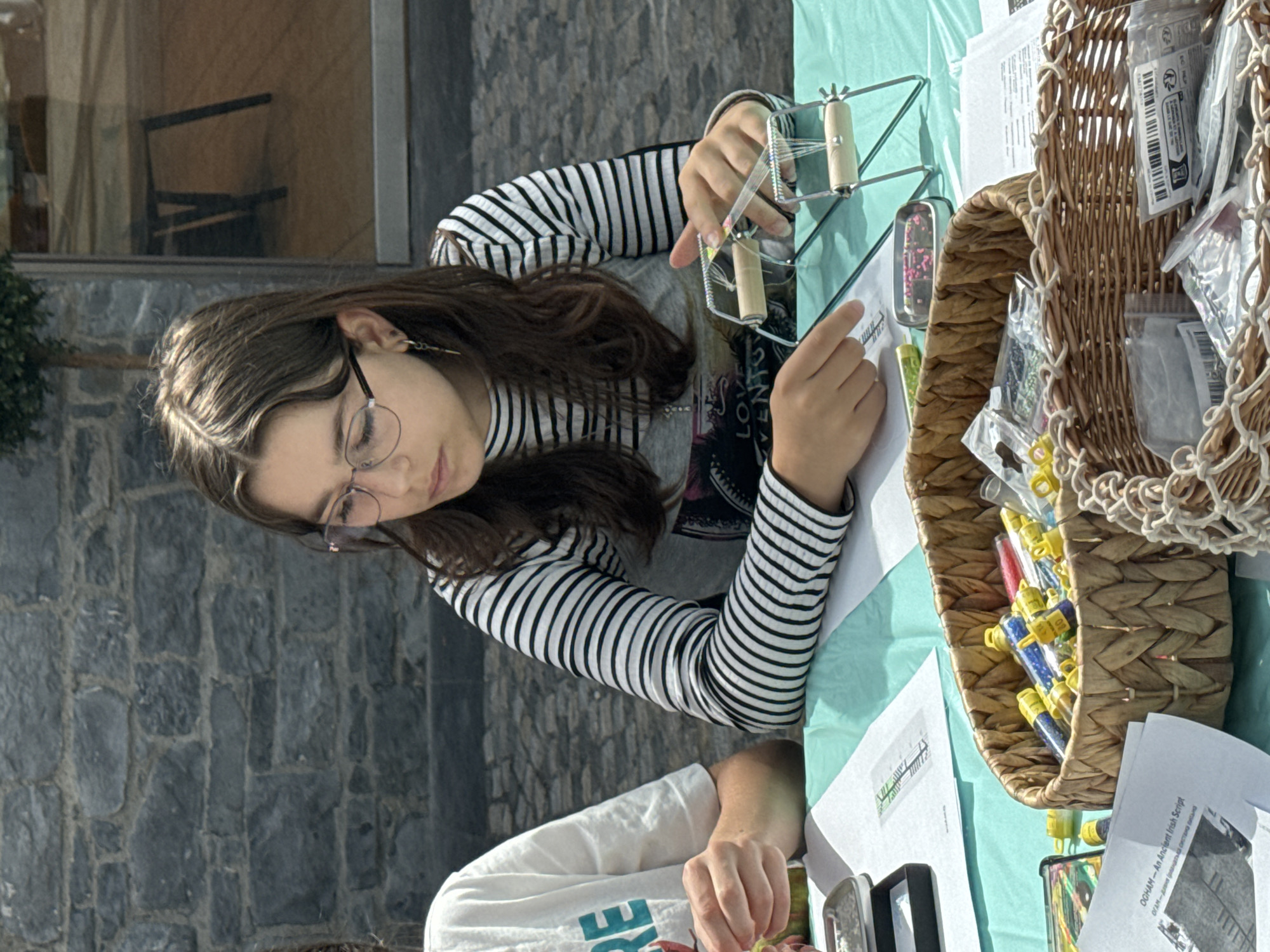 The image shows a young girl, wearing glasses and a striped long-sleeved shirt, sitting at a table covered with a teal tablecloth. She is engaged in a craft activity, specifically using a beading loom to create a piece of jewelry or craft. Her hands are actively manipulating threads and beads. In front of her, there is a woven basket overflowing with small vials of colorful beads, some loose beads, and other craft supplies. Several pieces of paper with text and diagrams are also visible on the table, one of which has the title "OGHAM - An Ancient Irish Script". The background features a stone wall and a glass-fronted building, suggesting an outdoor or semi-outdoor setting. The scene is likely taking place in the city of Kilkenny, Ireland, possibly at a craft fair, market, or educational event related to Irish heritage.