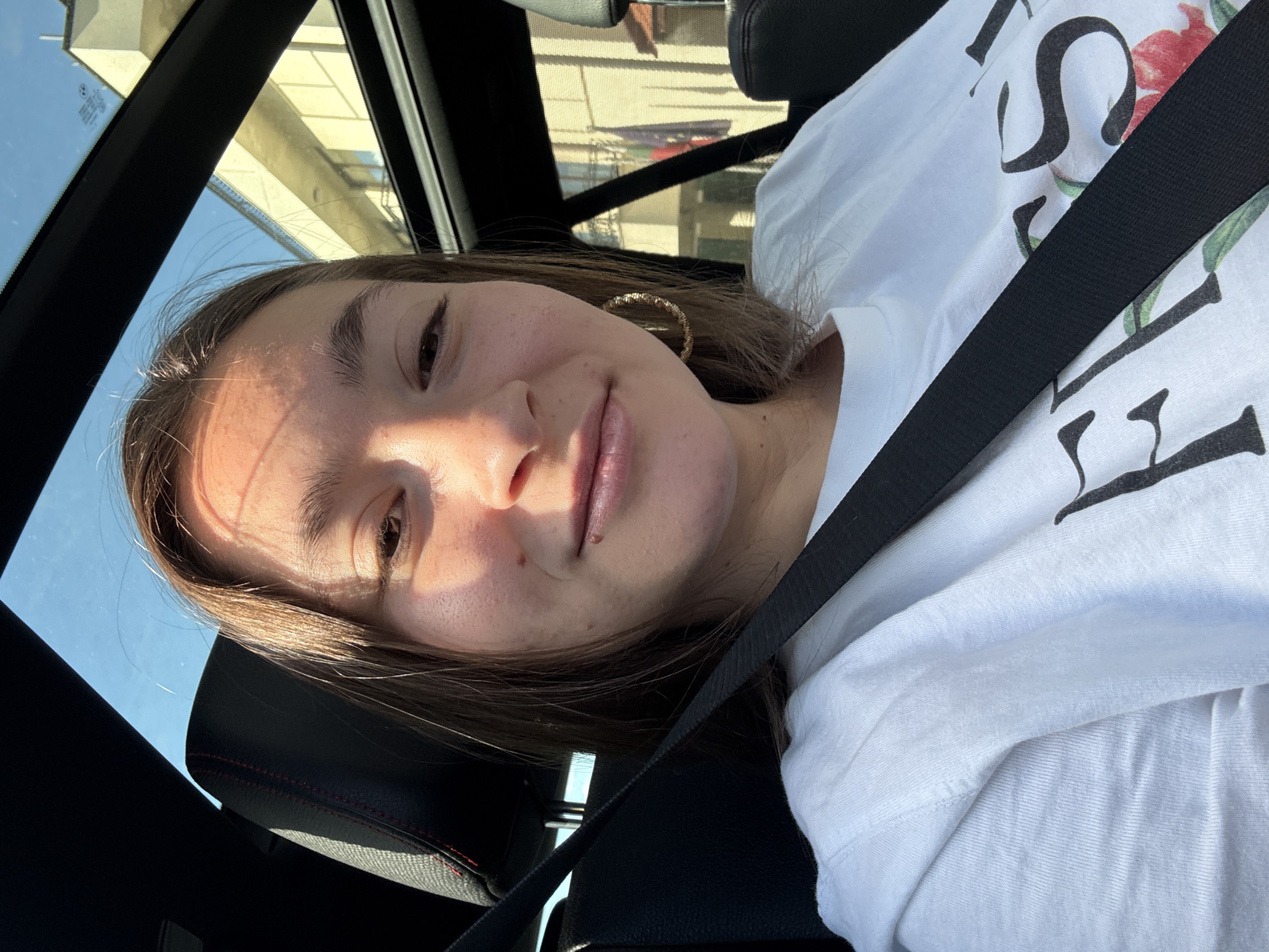 A young woman with light brown hair is taking a selfie from the passenger seat of a car. She is wearing a white t-shirt with a floral design and text that appears to say "FASHION". She has on large hoop earrings. The car has a sunroof, and sunlight is streaming through it, casting a shadow across her face. The interior of the car is visible, including a black seat and a seatbelt. Outside the car, a building with yellow siding and balconies is visible. The location is the city of Sofia, Bulgaria.