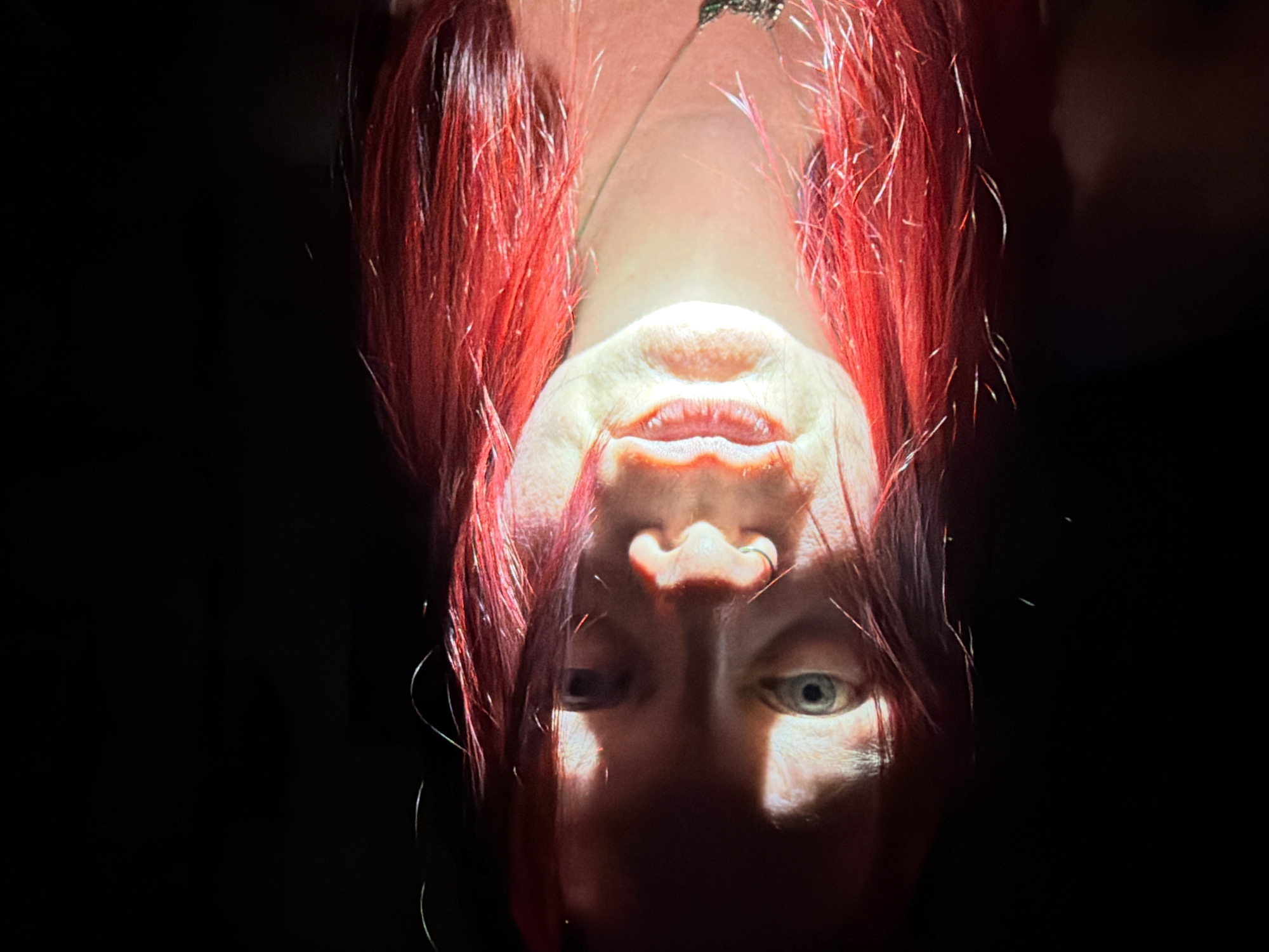 An upside-down, close-up portrait of a person with red hair is dominated by stark lighting that illuminates the mouth and nose. The rest of the face is partially shrouded in shadow. One visible eye is light-colored. A necklace is visible at the top of the image. The background is dark and indistinct. There is no evidence of location in Chaikovsky, Russia.