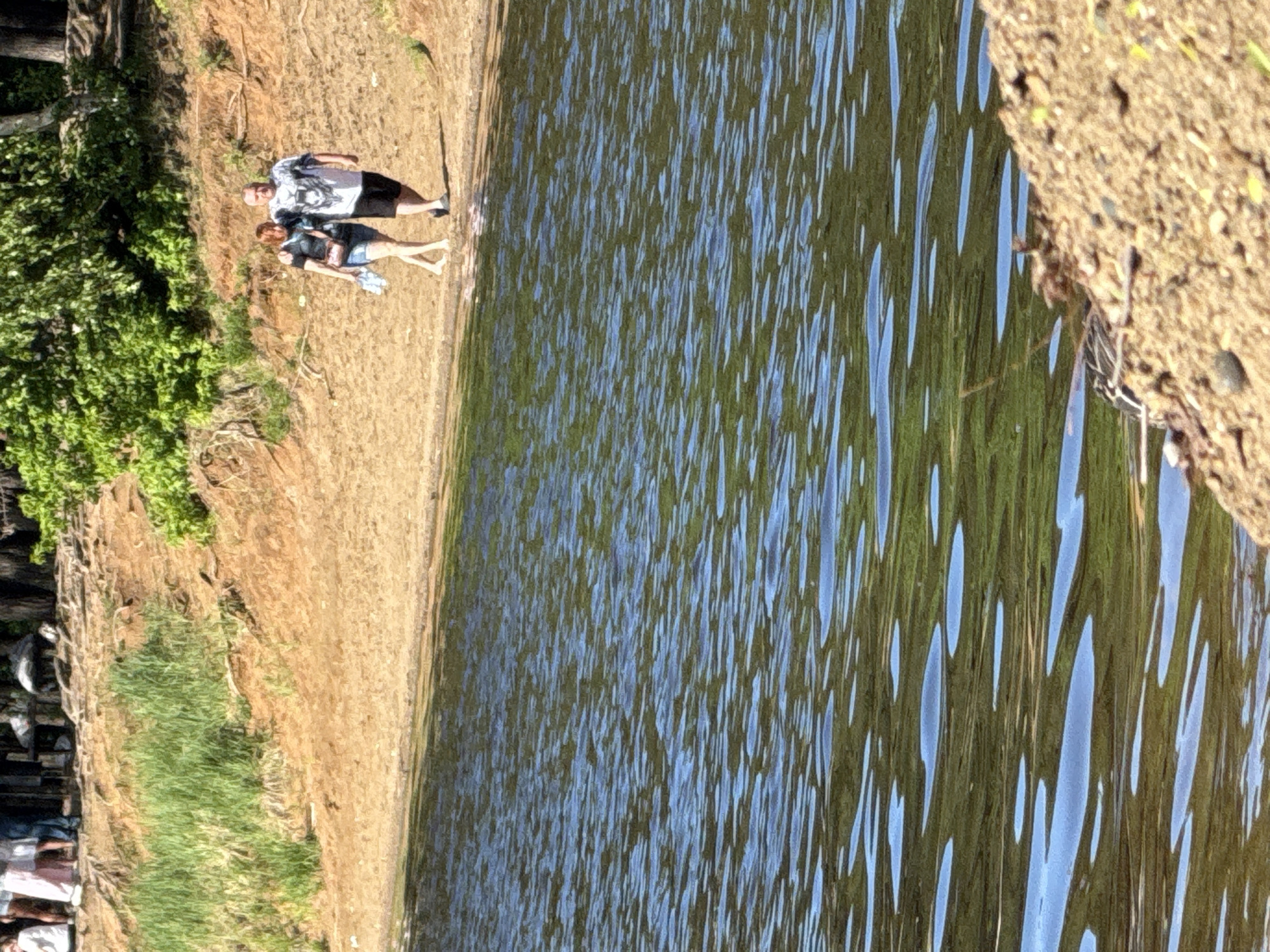 The image depicts a body of water, likely a lake or river, with rippling surfaces reflecting the sky. The shoreline is visible, consisting of sandy soil and some vegetation. On the shore, two individuals stand together. One appears to be male wearing a t-shirt and shorts. Next to him is a female with an arm around him, wearing a t-shirt and shorts. Based on the scene and given location, it is plausible this is a view of the Kama River. The context of their presence and actions requires further information.