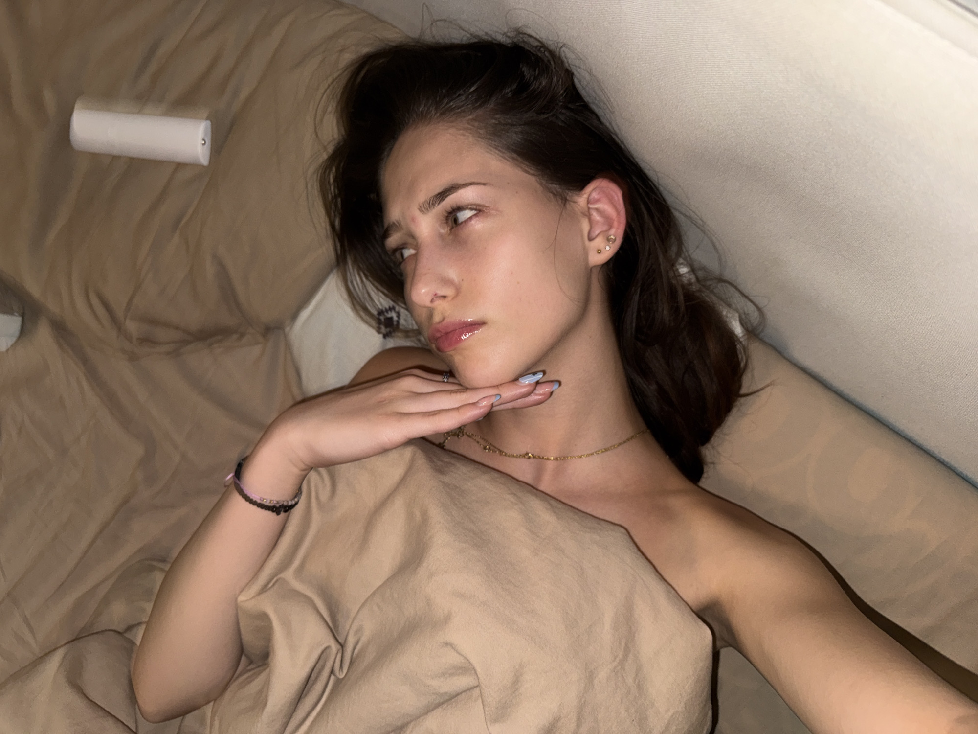 A young woman is lying on a bed, partially covered by a tan blanket. Her head is tilted back and her eyes are looking upwards. She is wearing a gold necklace and bracelets on her left wrist. Her nails are painted light blue. There are two small earrings in her right ear. A white cylindrical object, possibly a remote or a small flashlight, is visible on the bed in the upper right corner of the frame. The bed appears to be in a room with tan and beige bedding. The image appears to be taken indoors, in what could be a bedroom. There is no information in the image to suggest a specific location in the city of Sofia, Bulgaria.