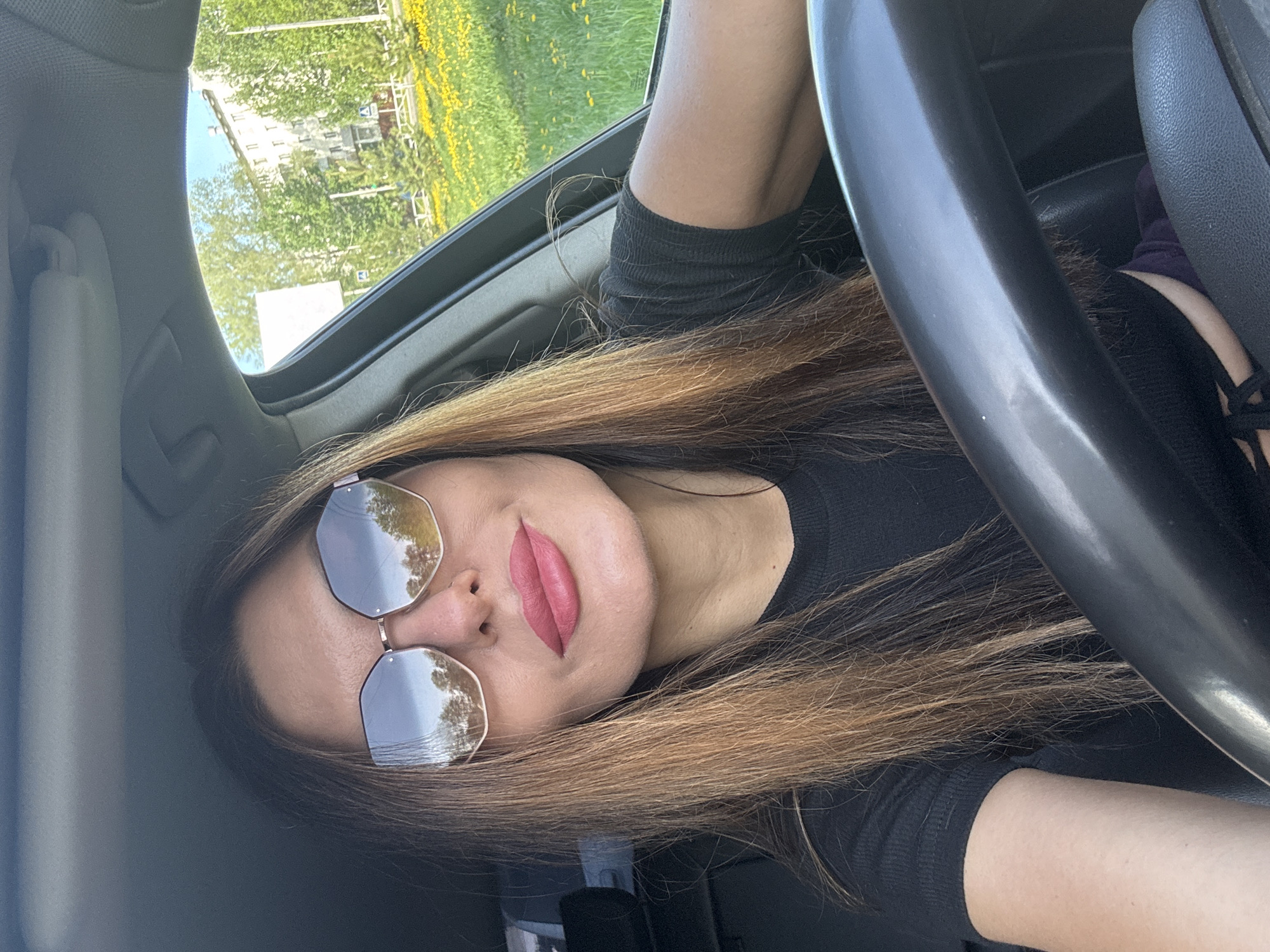 A woman is seated in a car. She has light brown hair and is wearing sunglasses. Her lips are a dark pink color. A steering wheel is visible in the frame. Outside the window, trees and a building can be seen. The location is Chaikovsky, Russia.