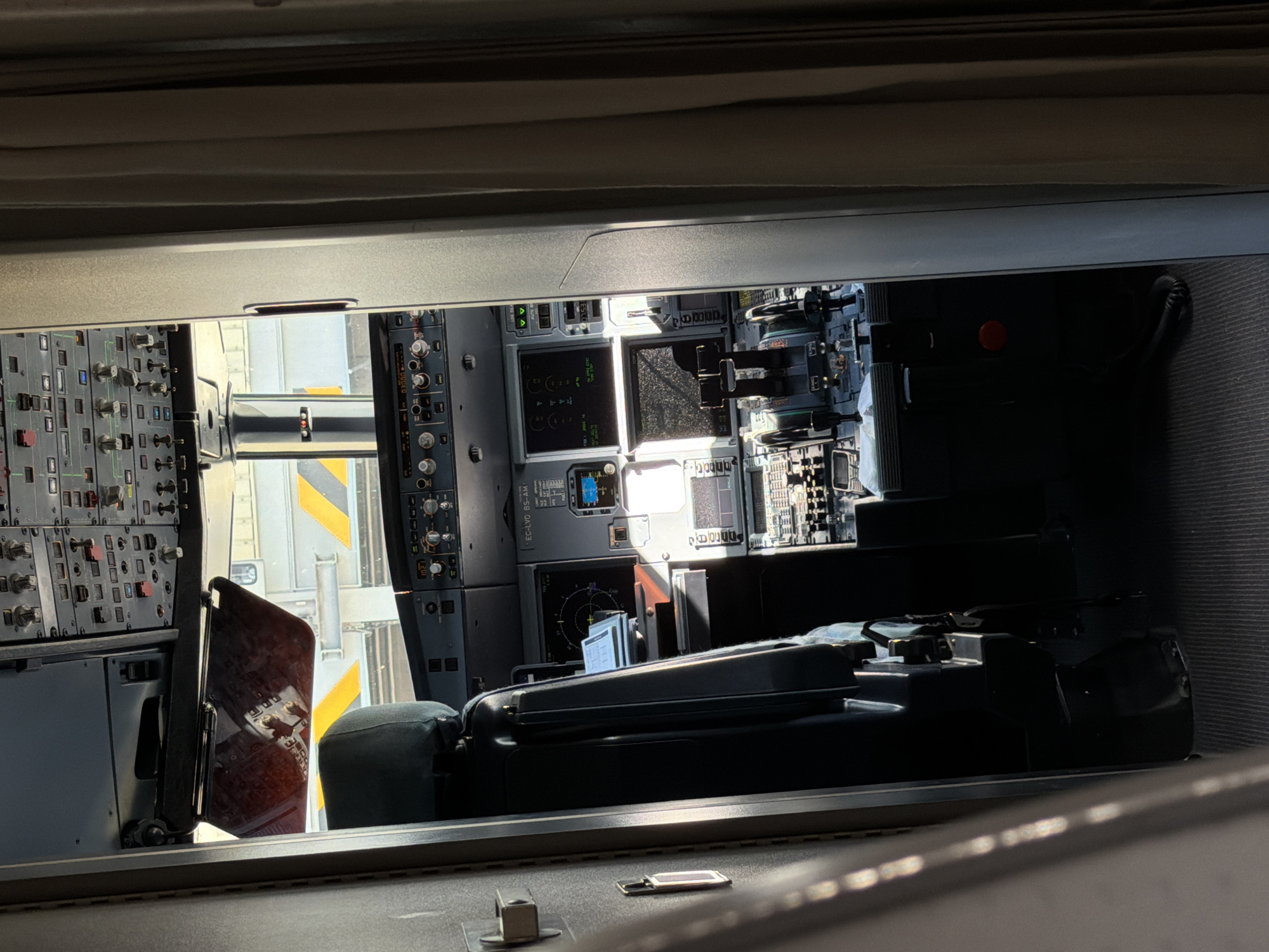 This image provides an interior view of a modern aircraft cockpit, seen from an elevated position, likely from a cabin area or observation window. The scene is devoid of people but filled with an extensive array of aviation technology.
The primary subjects are the complex control panels and displays of a commercial airliner. The main instrument panel is densely packed with numerous buttons, switches, dials, and illuminated digital screens. Key flight displays are visible, showing various flight parameters and navigational data. One screen clearly displays the aircraft registration "EC-LVO BS-AM," indicating a Spanish-registered aircraft. Below the main panel, a central pedestal houses what appear to be thrust levers and other operational controls. Dark upholstered pilot seats are positioned in front of the controls.
Through the cockpit windows, an exterior scene is visible, suggesting the aircraft is on the ground at an airport. Bright daylight illuminates the area, indicating clear weather. Ground equipment, possibly part of a jet bridge or service vehicle, can be seen with distinctive yellow and black striped safety markings.
No activity involving people is taking place; the cockpit appears to be powered on, as indicated by the illuminated displays and lights, but unoccupied. The overall impression is one of technical sophistication and readiness. The detailed crops enhance the visibility of individual controls and screens, emphasizing the intricate design of the cockpit environment. The registration "EC-LVO" suggests the aircraft is registered in Spain, and given the context of Crawley, United Kingdom, the photograph was likely taken at a nearby international airport such as London Gatwick.