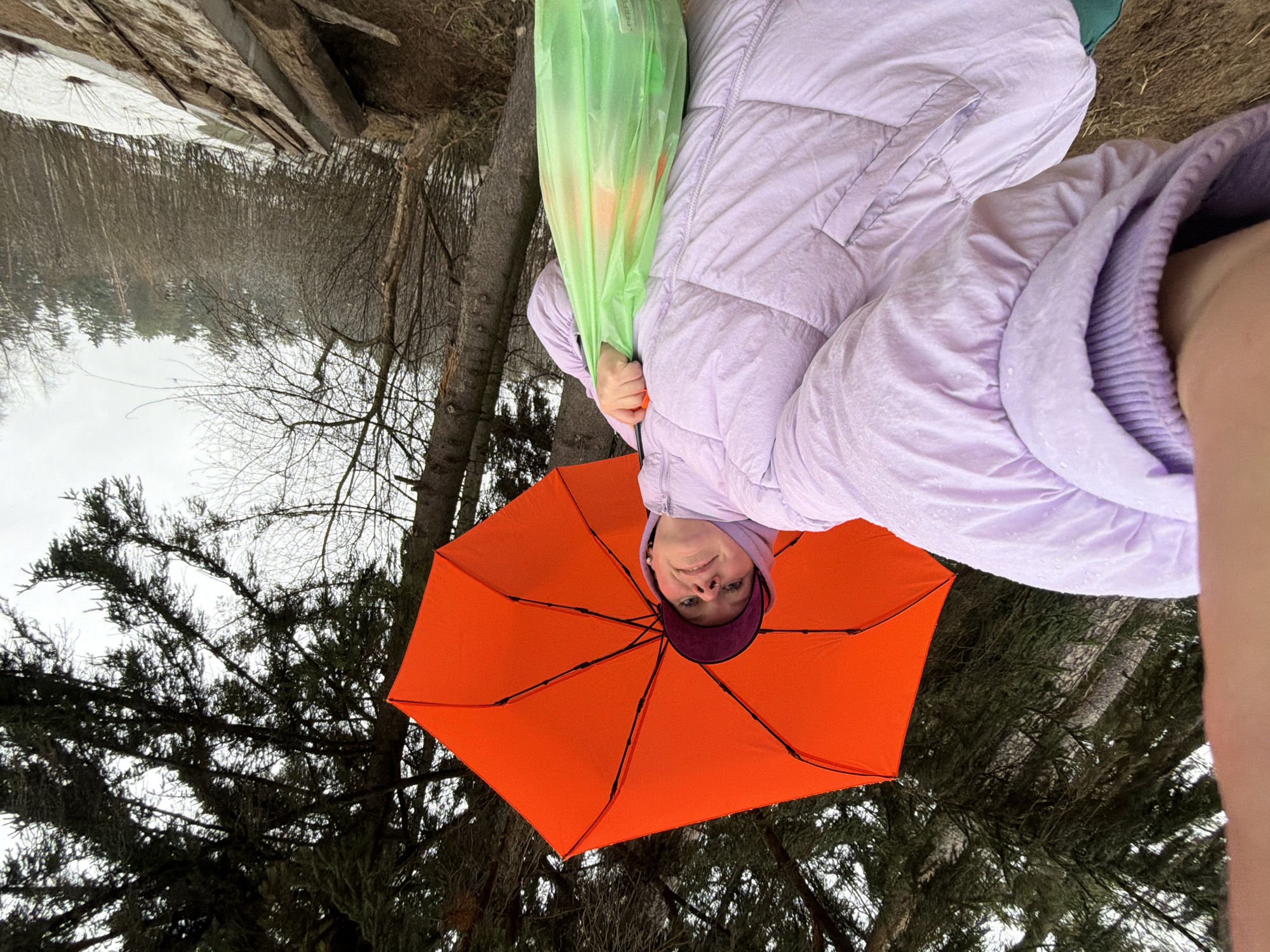 A person is holding an orange umbrella, viewed from below, with trees and a body of water in the background. The person wears a light purple puffer jacket and a dark purple cap. The location is possibly near Chaikovsky, Russia, suggested by elements such as the type of vegetation and wintery conditions, though precise city verification demands more data. The view direction is primarily focused on the person and the umbrella, with the natural environment providing a backdrop of trees and water under overcast weather conditions.