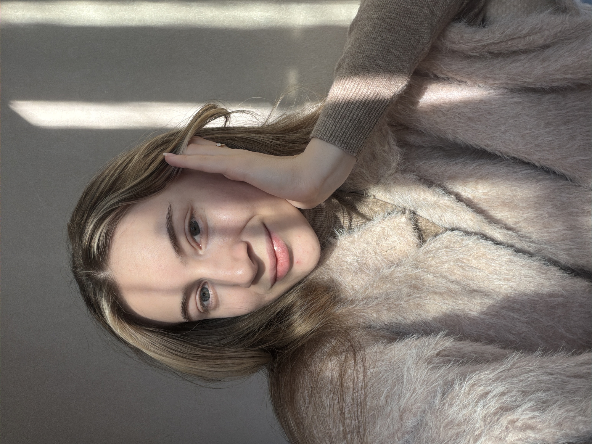 A fair-skinned woman with light brown hair, streaked with blonde highlights, poses indoors. Her left hand is raised to her face, with the fingers near her forehead and the palm against her cheek. She has light-colored eyes, a small nose, and her lips are slightly parted in a smile. She is wearing a light brown sweater and a fluffy light brown coat, suggesting it might be cold. Strong sunlight casts a sharp shadow across the upper left of the image and onto the wall in the background. There's no information to infer a specific location within Plovdiv, Bulgaria, or to confidently place the scene in that city at all.